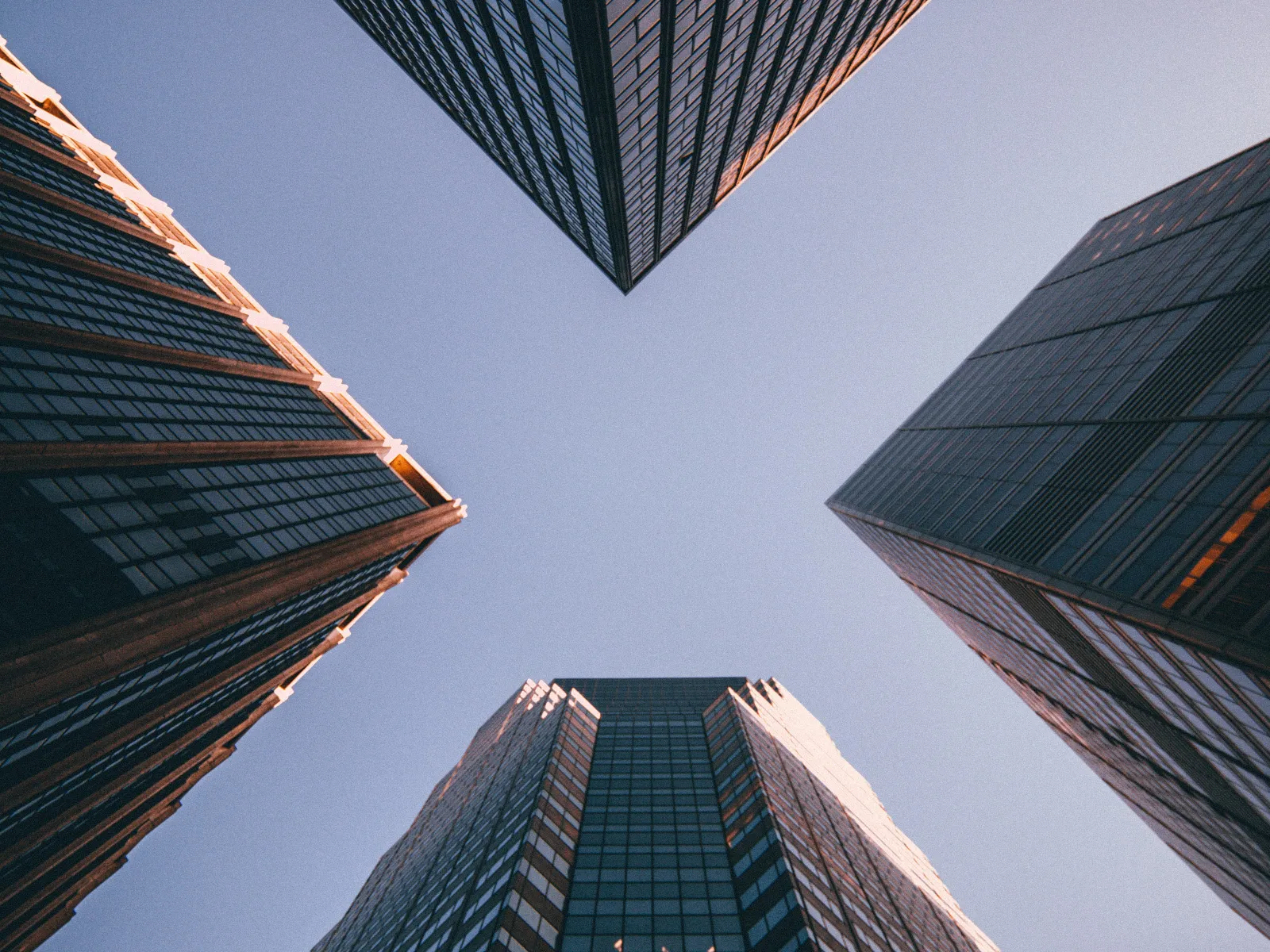 Looking up at glass skyscrapers converging under a clear blue sky reflecting golden sunlight in urban downtown.