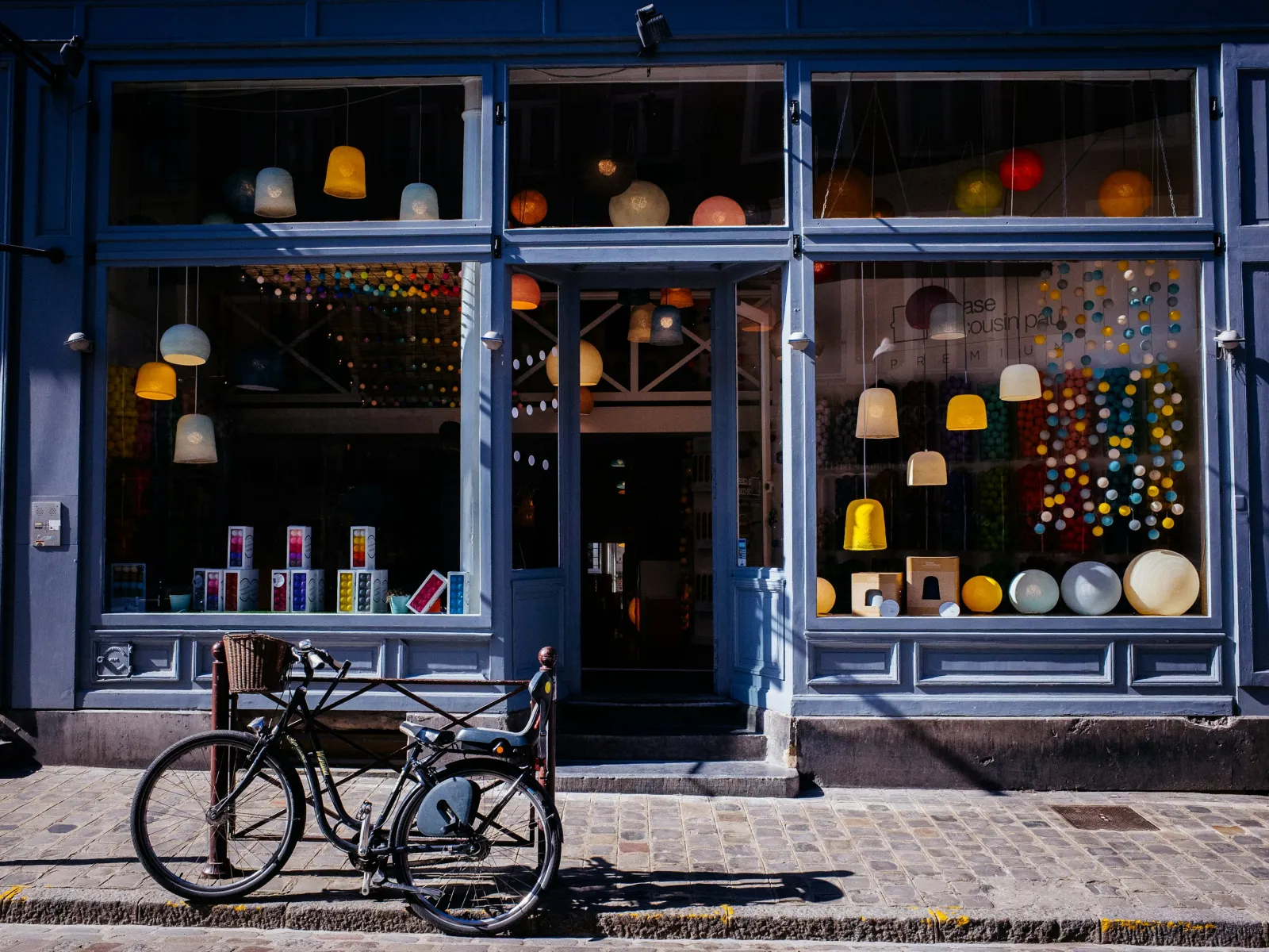 Blue storefront with hanging lamps and colorful decorations reflected in glass windows, bicycle parked outside on cobblestone street