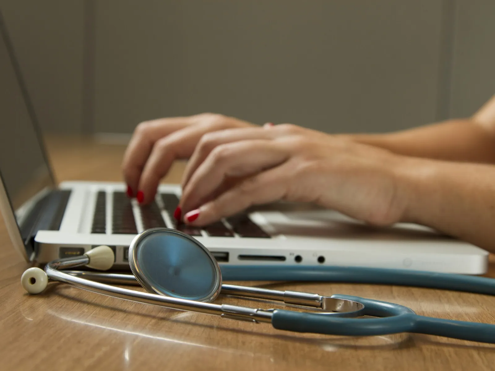 Hands typing on a laptop keyboard next to a stethoscope on a wooden table, symbolizing medical work.