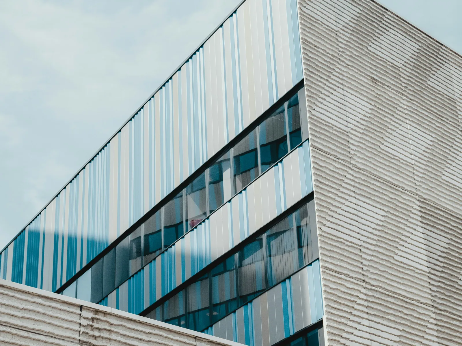 Modern building with textured beige bricks and reflective vertical blue glass windows under a cloudy sky.