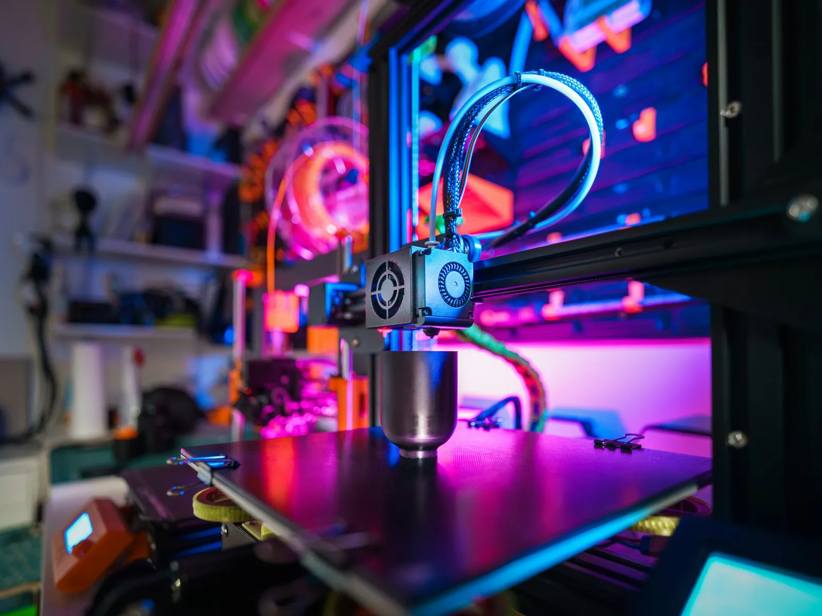 Close-up of a 3D printer nozzle and heated bed with colorful LED lighting in a tech workspace.