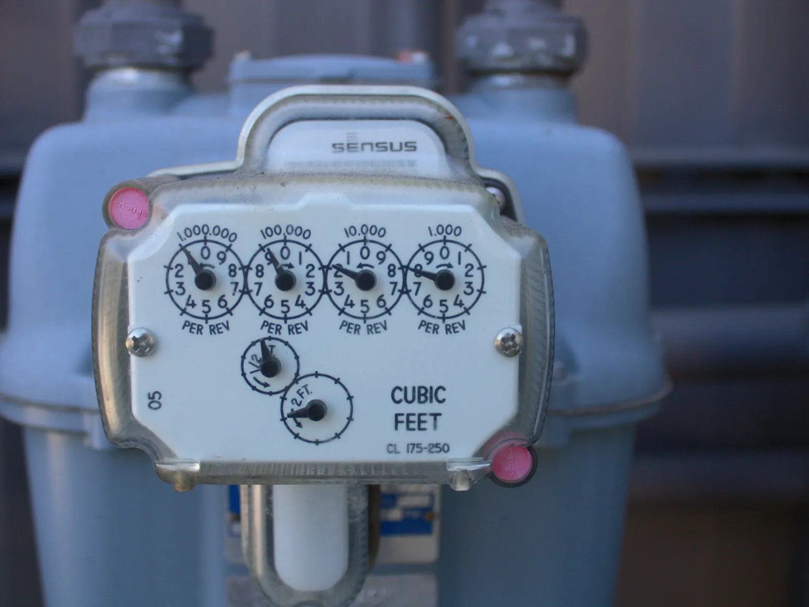 Close-up of a Sensus gas meter showing dials measuring cubic feet with industrial pipes in the background