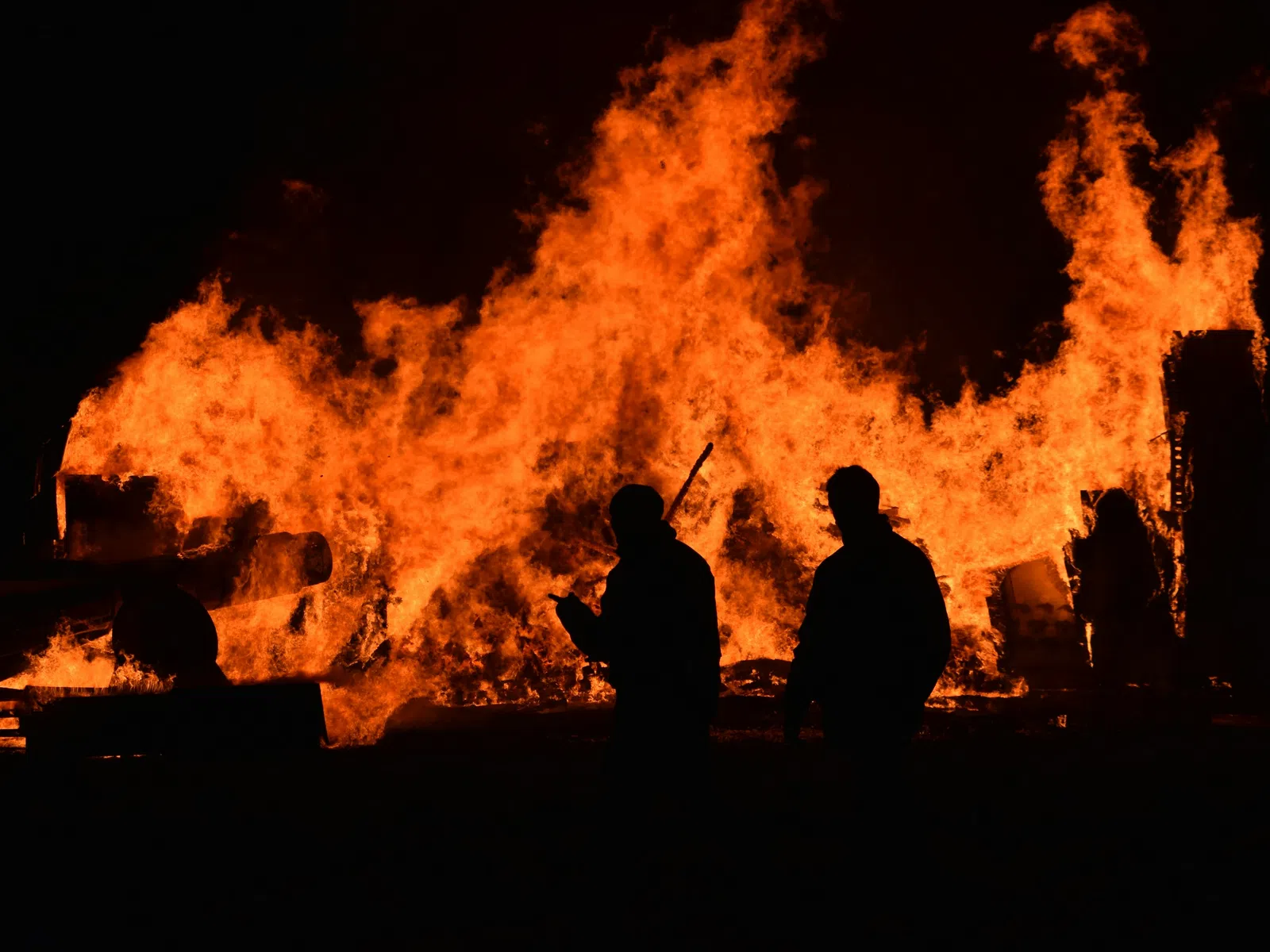 Silhouettes of two people standing near a large raging fire at night with intense orange flames.