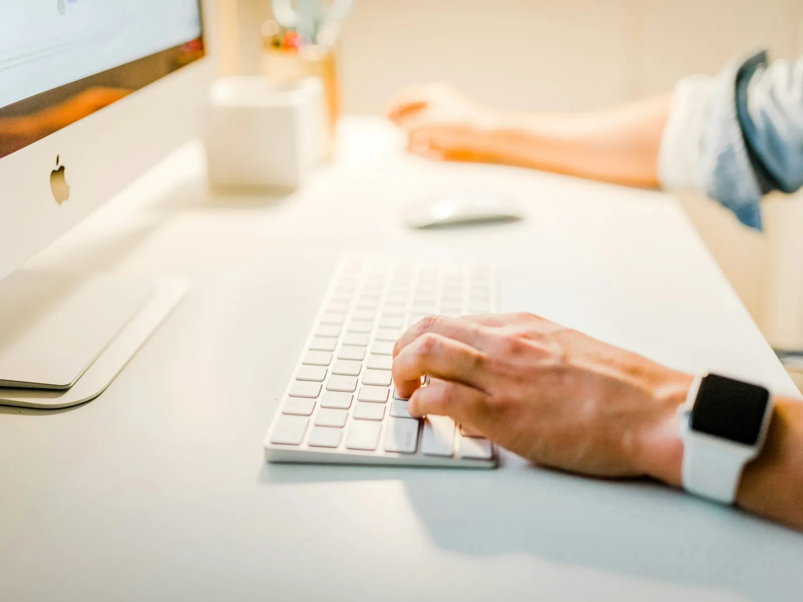 Close-up of a person typing on a white keyboard beside an Apple desktop computer on a clean desk.