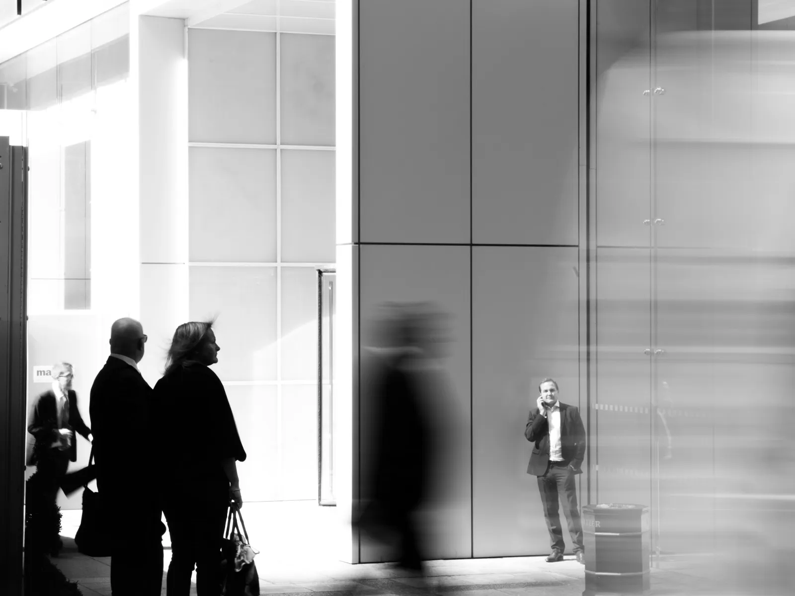 Black and white photo of people walking and standing near a modern building with reflective glass panels.