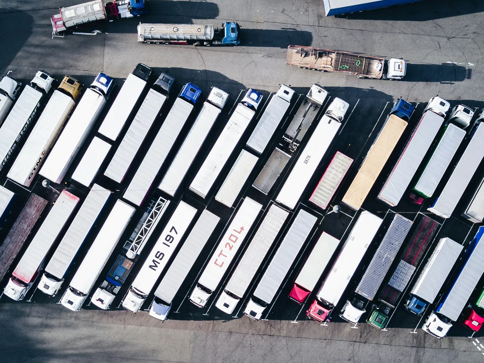 Aerial view of numerous parked semi trucks and trailers in a logistics parking lot on a sunny day.