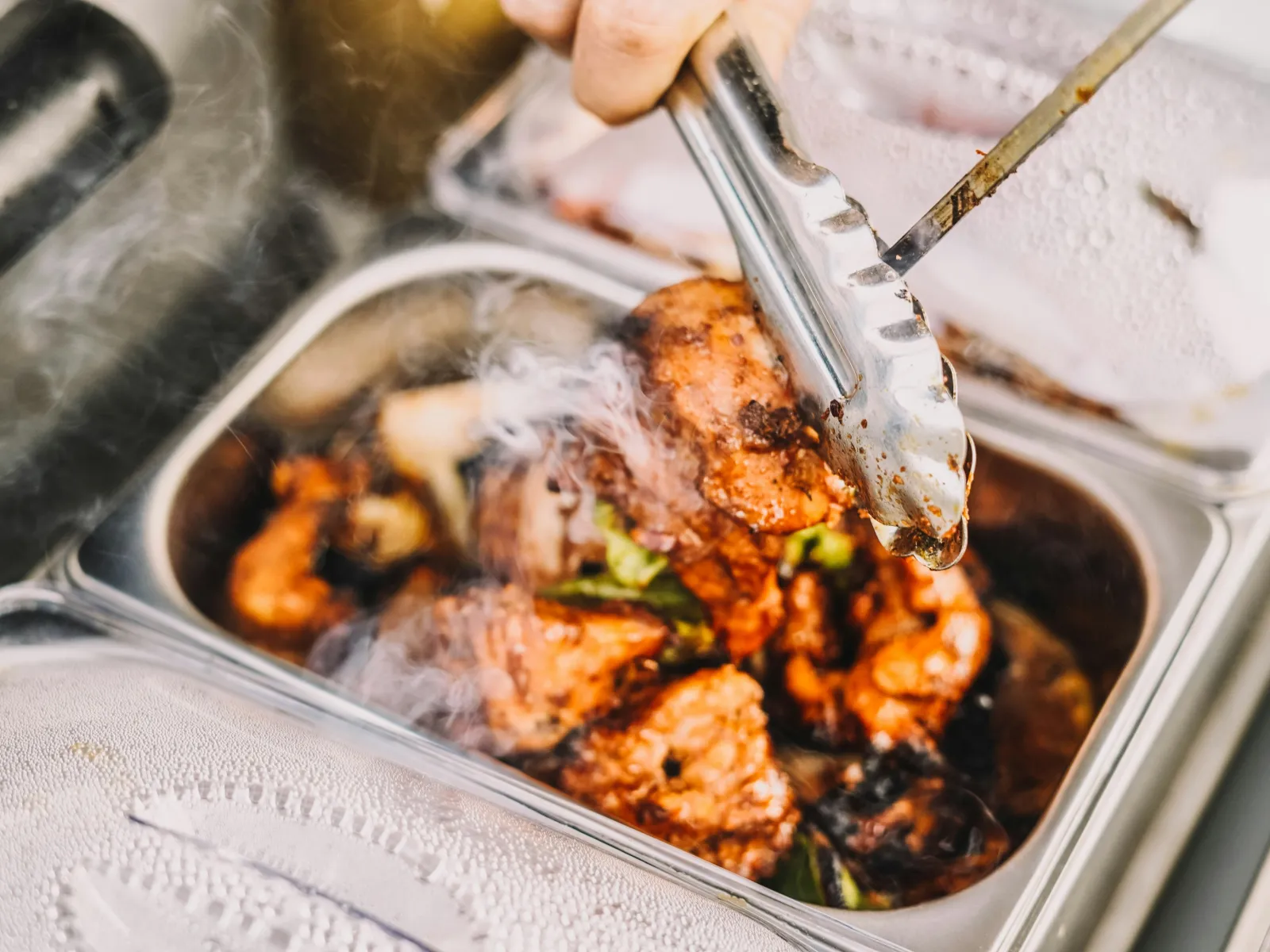 Hand holding tongs picking up steaming grilled or roasted spicy chicken pieces from a metal tray.