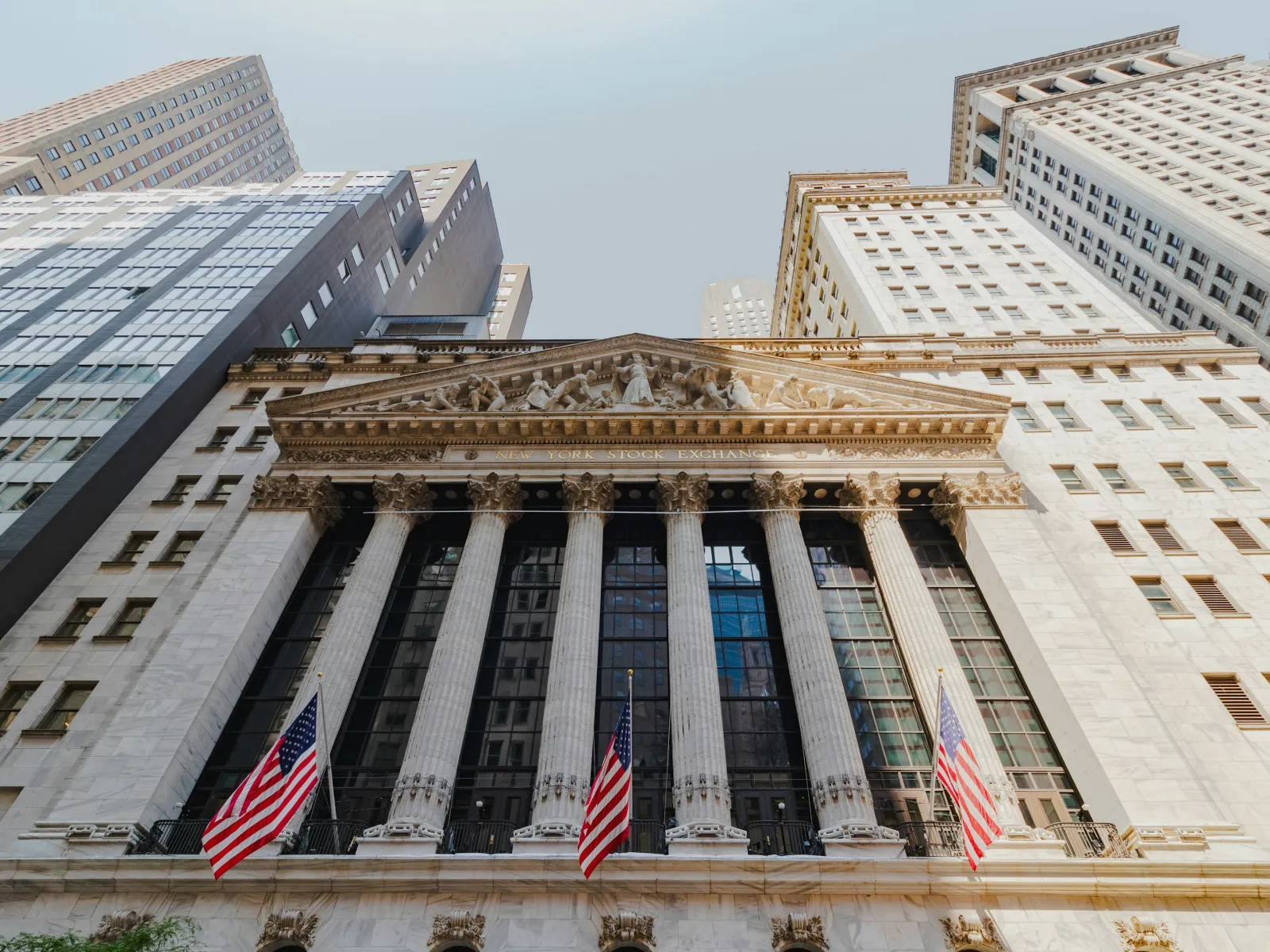 Facade of New York Stock Exchange building with American flags and surrounding skyscrapers under clear sky.