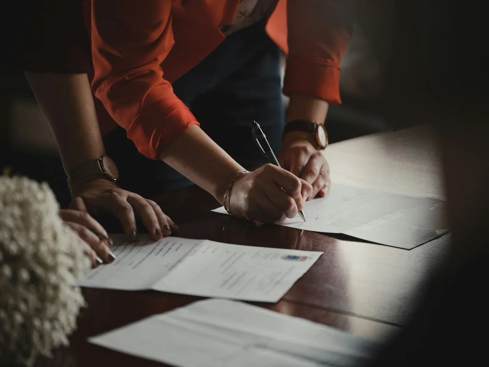 Person in orange jacket signing documents on a wooden table with another person nearby reviewing papers.