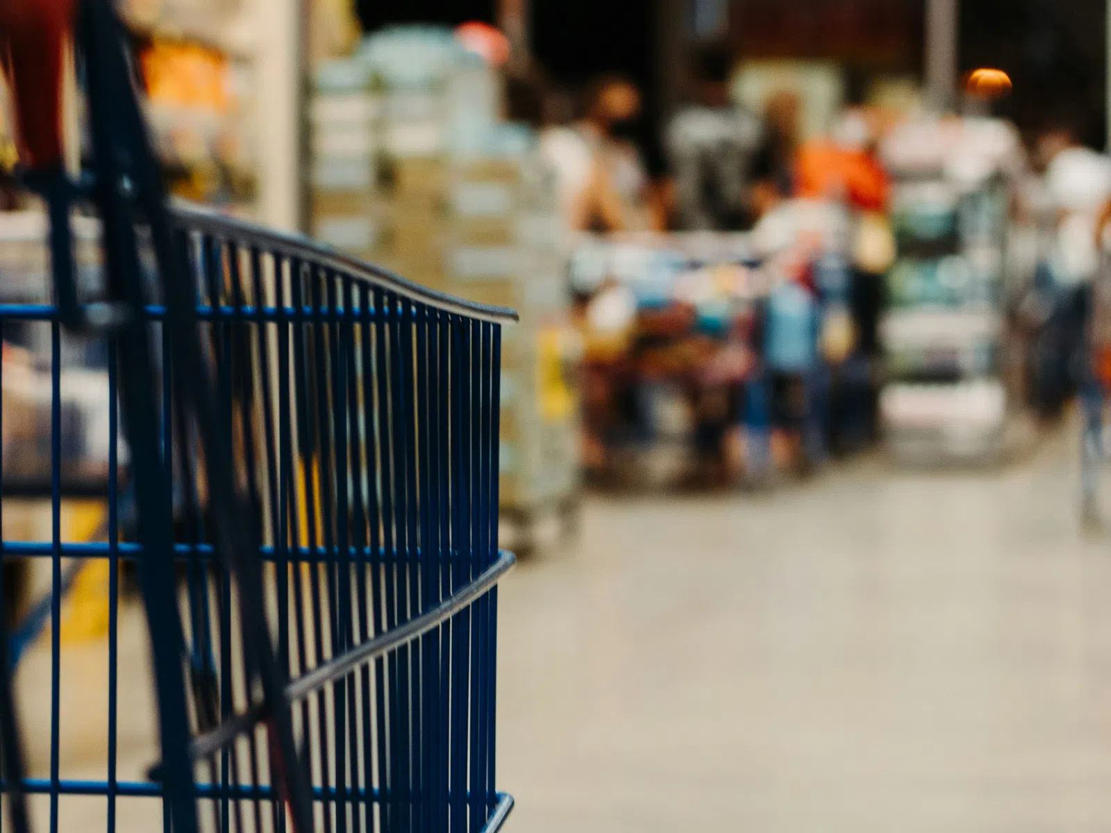 Empty blue shopping cart in aisle of warehouse store with blurred shoppers and products in background