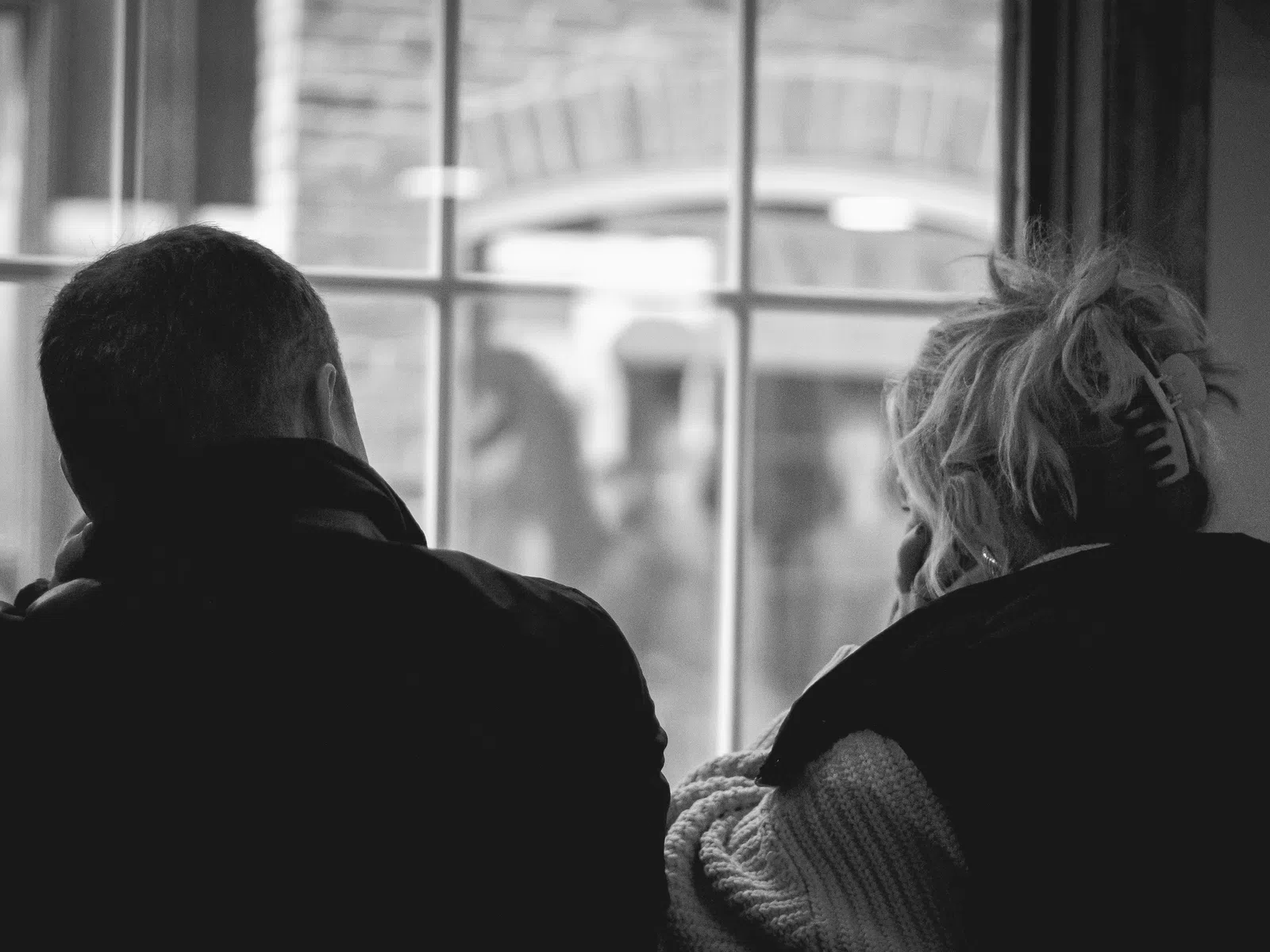 Black and white photo of two people sitting by a window, viewed from behind, in a cozy indoor setting.