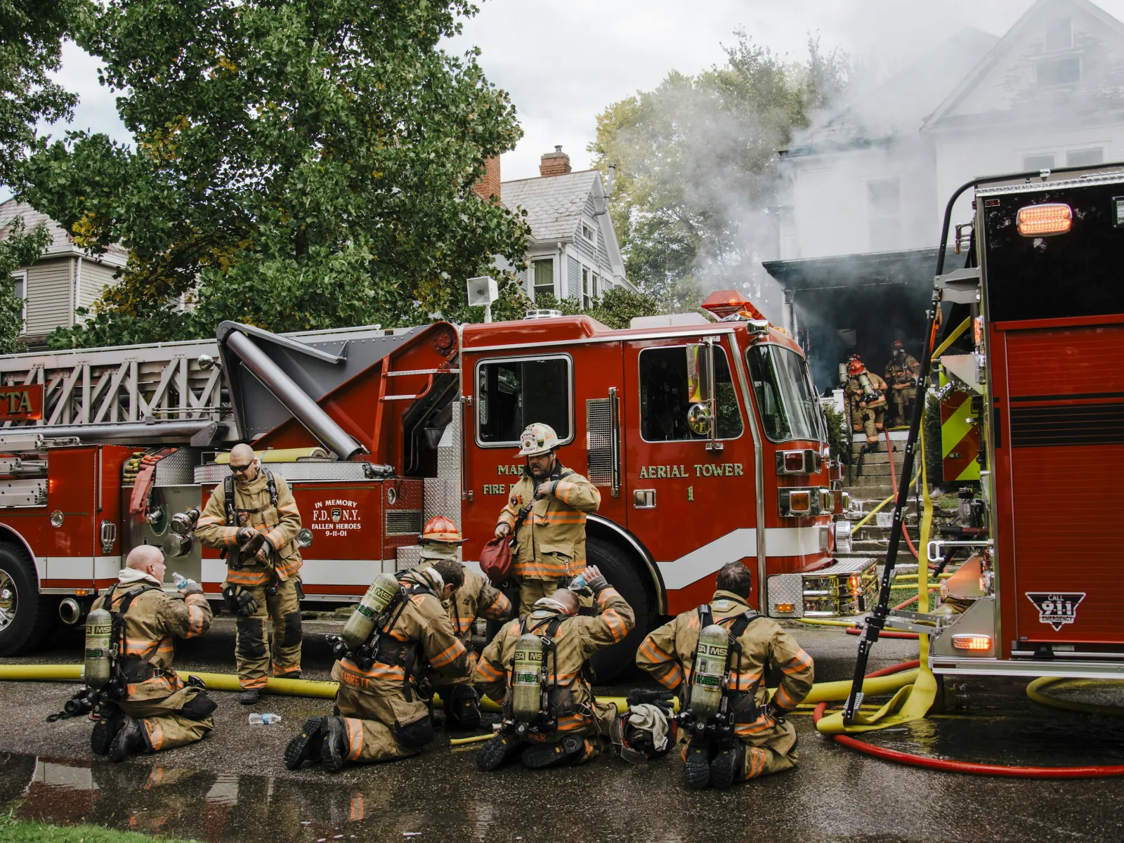 Firefighters in gear resting near a Marietta aerial tower fire truck at a smoky house fire scene.