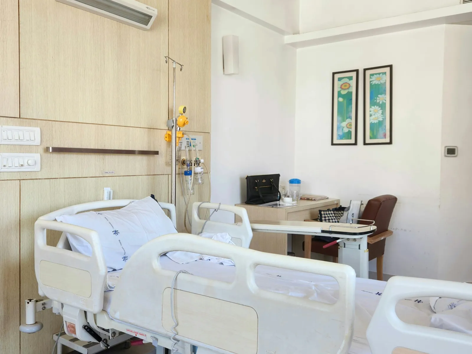 Clean hospital room with an adjustable bed, IV stand, wooden wall panel, and a desk with chair and decorations.