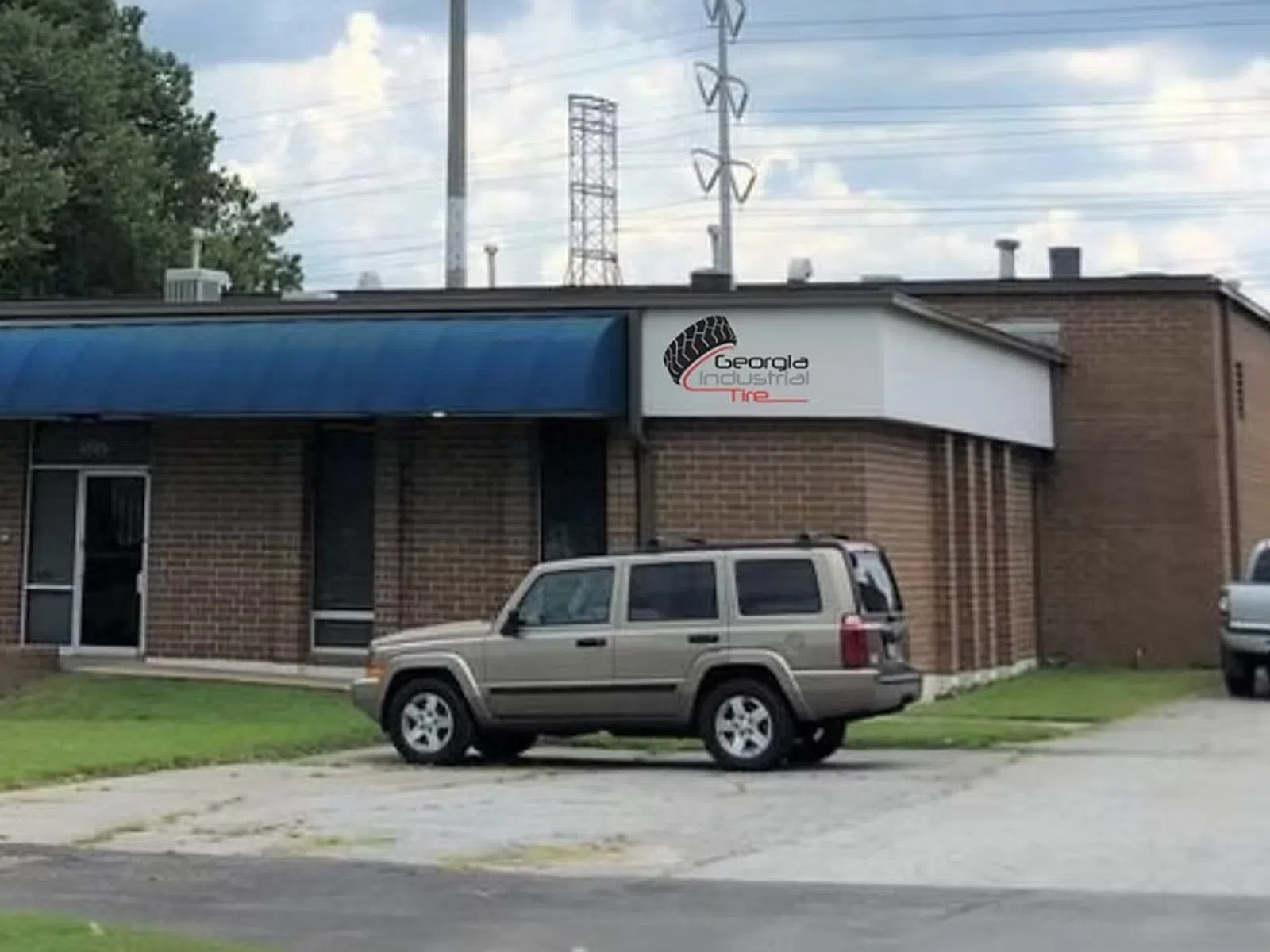 Brick office building with blue awning, parking lot with cars, and green lawn under partly cloudy sky.