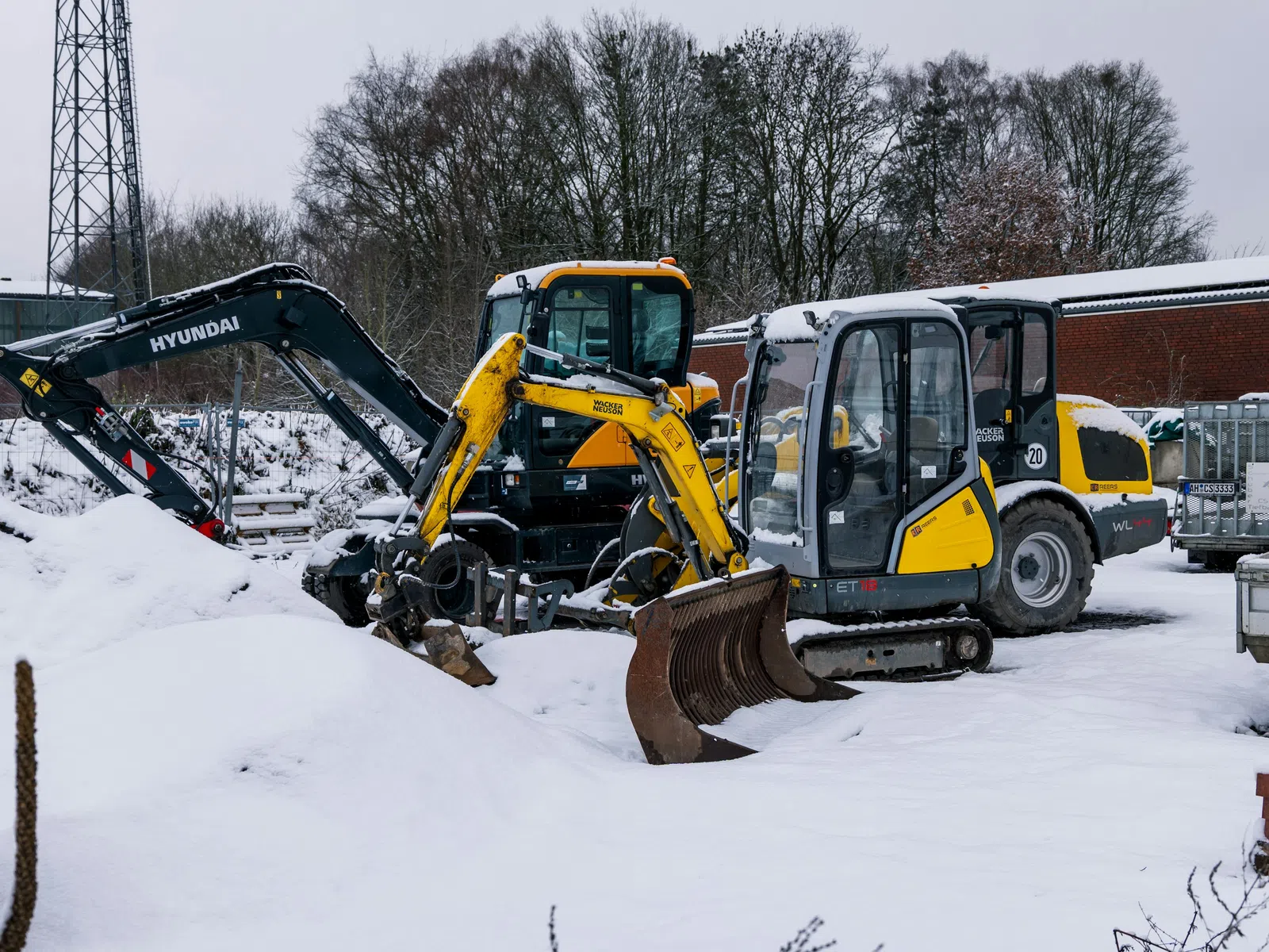 Yellow and black excavators parked on snow-covered ground near construction materials and bare trees in winter.