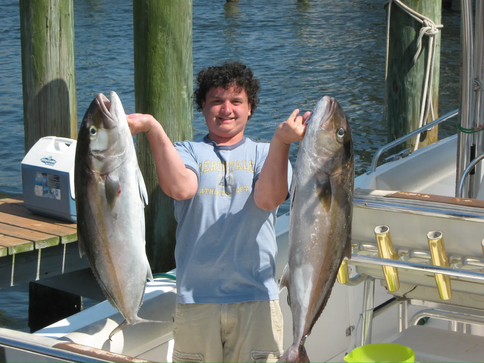 Man holding two large fish on a boat dock with water and wooden posts in the background.
