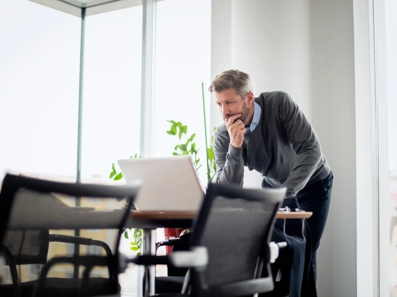 Man in sweater leaning over table working thoughtfully on laptop in bright office with large windows and plants.