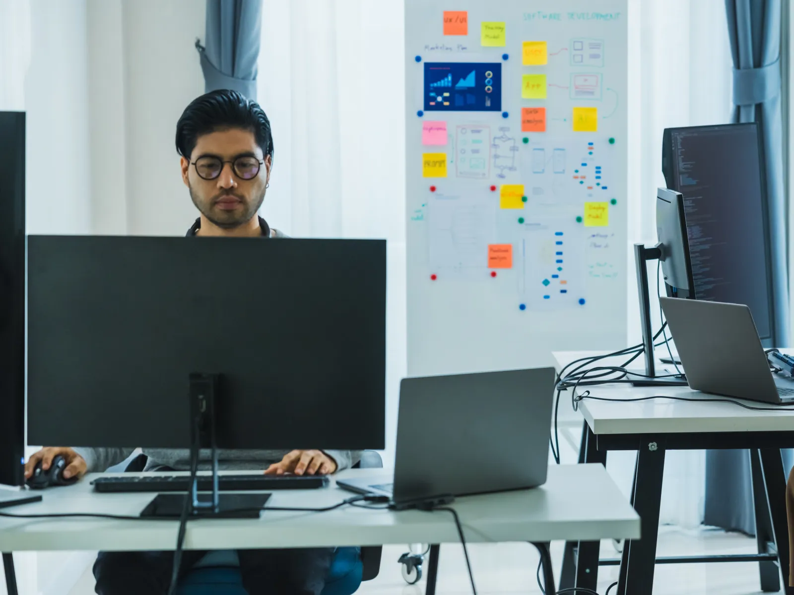 Two professionals working on computers in a modern office with charts and sticky notes on a whiteboard.