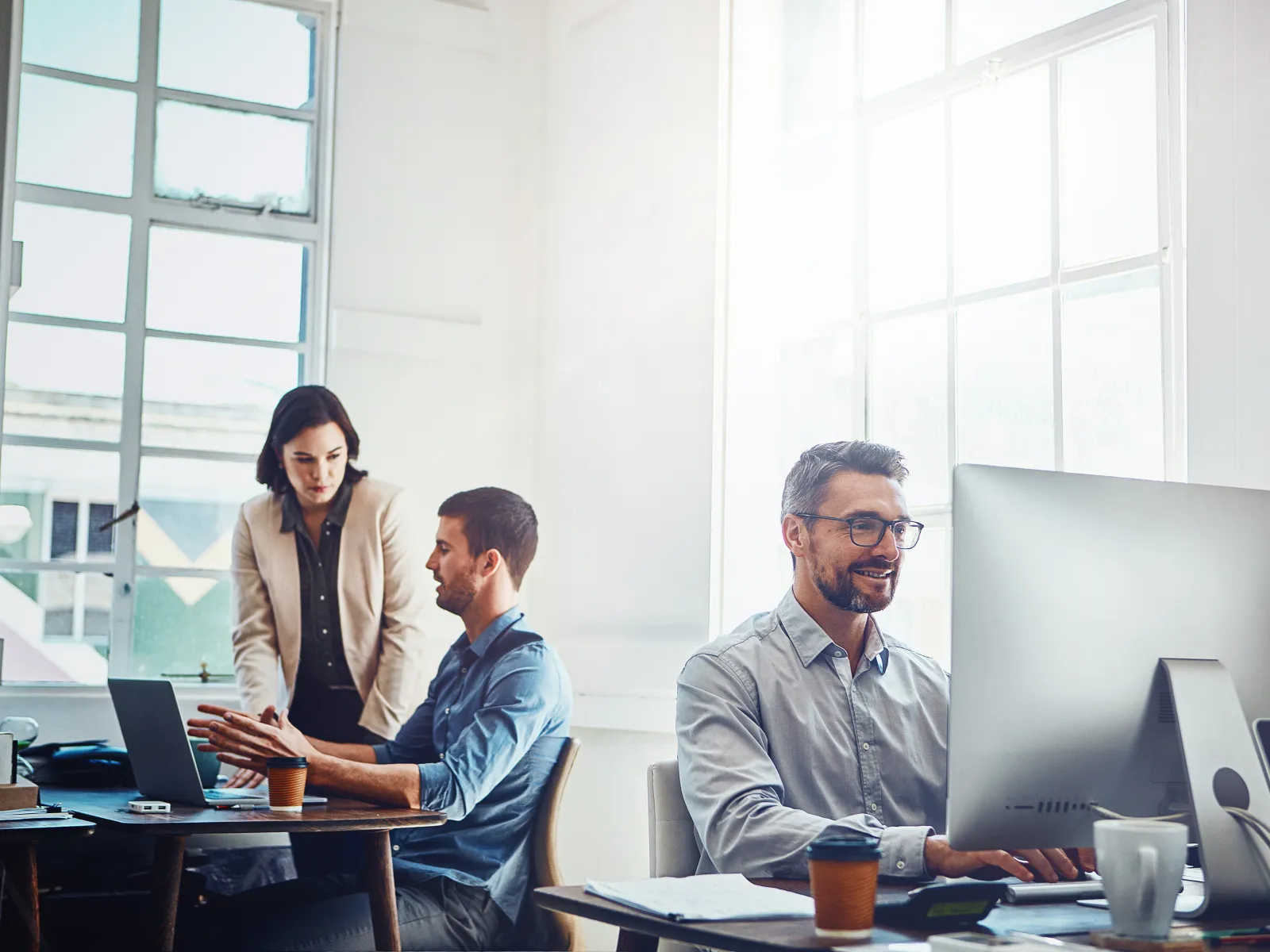 Three professionals working together in a bright modern office with computers and coffee cups on desks.