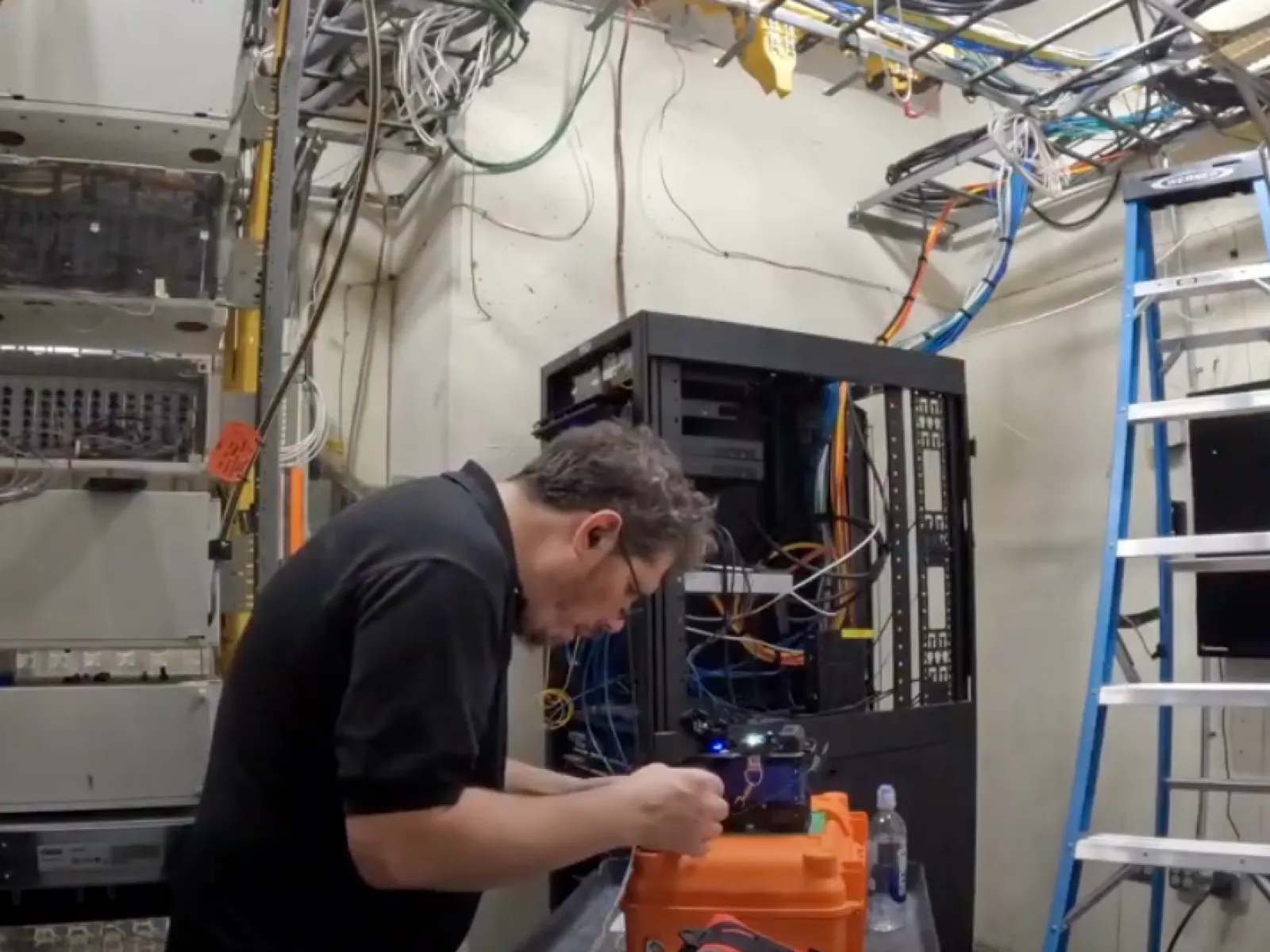 Technician working on fiber optics equipment in a server room with cables, racks, and a ladder.