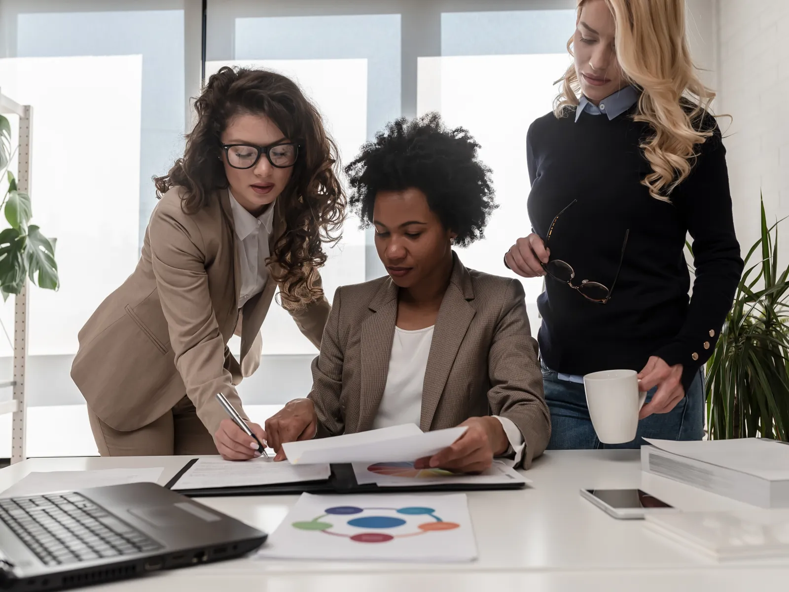 Three professional women collaborating over documents in a bright modern office space with laptop and coffee.
