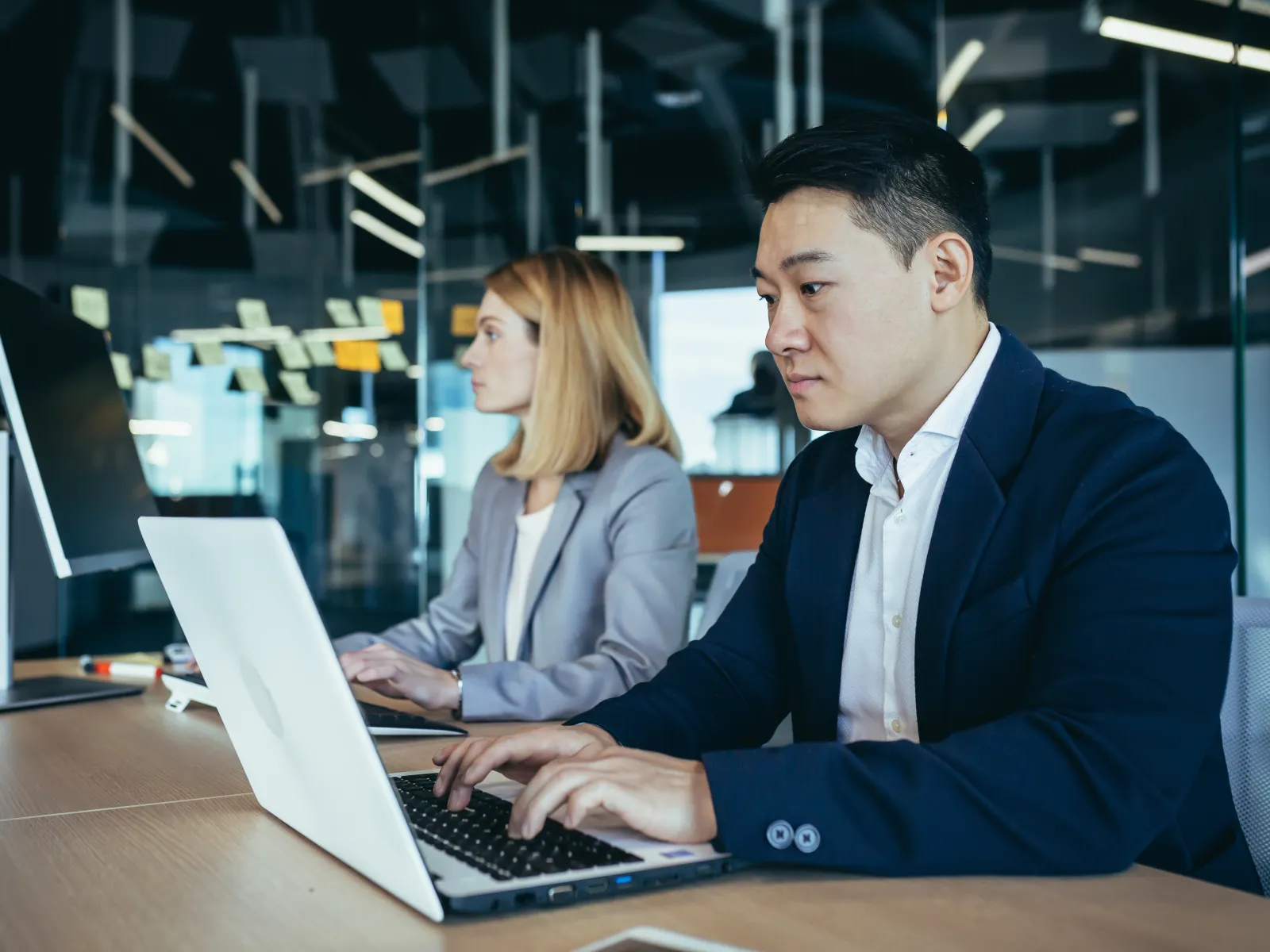 Two business professionals working on laptops in a modern office with glass walls and sticky notes.