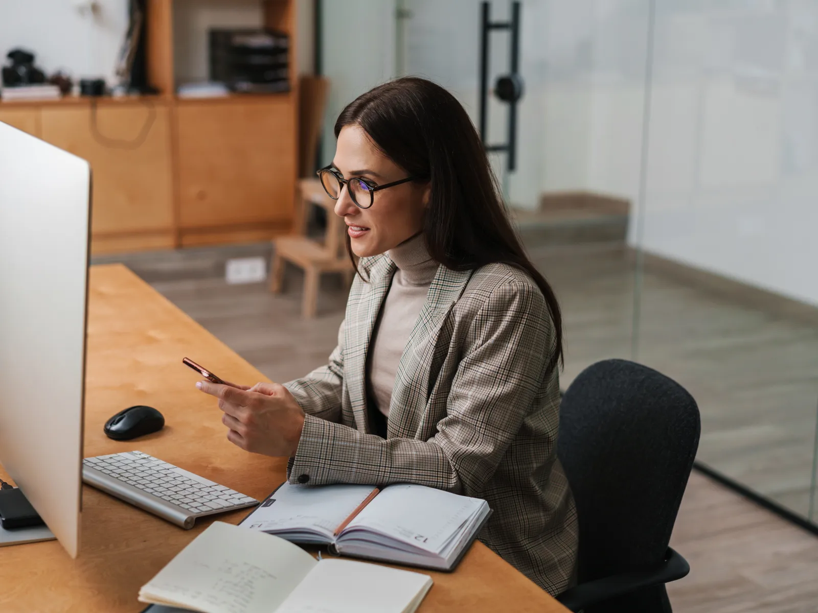 Woman in glasses working at desk with computer, smiling and using smartphone in modern office space.