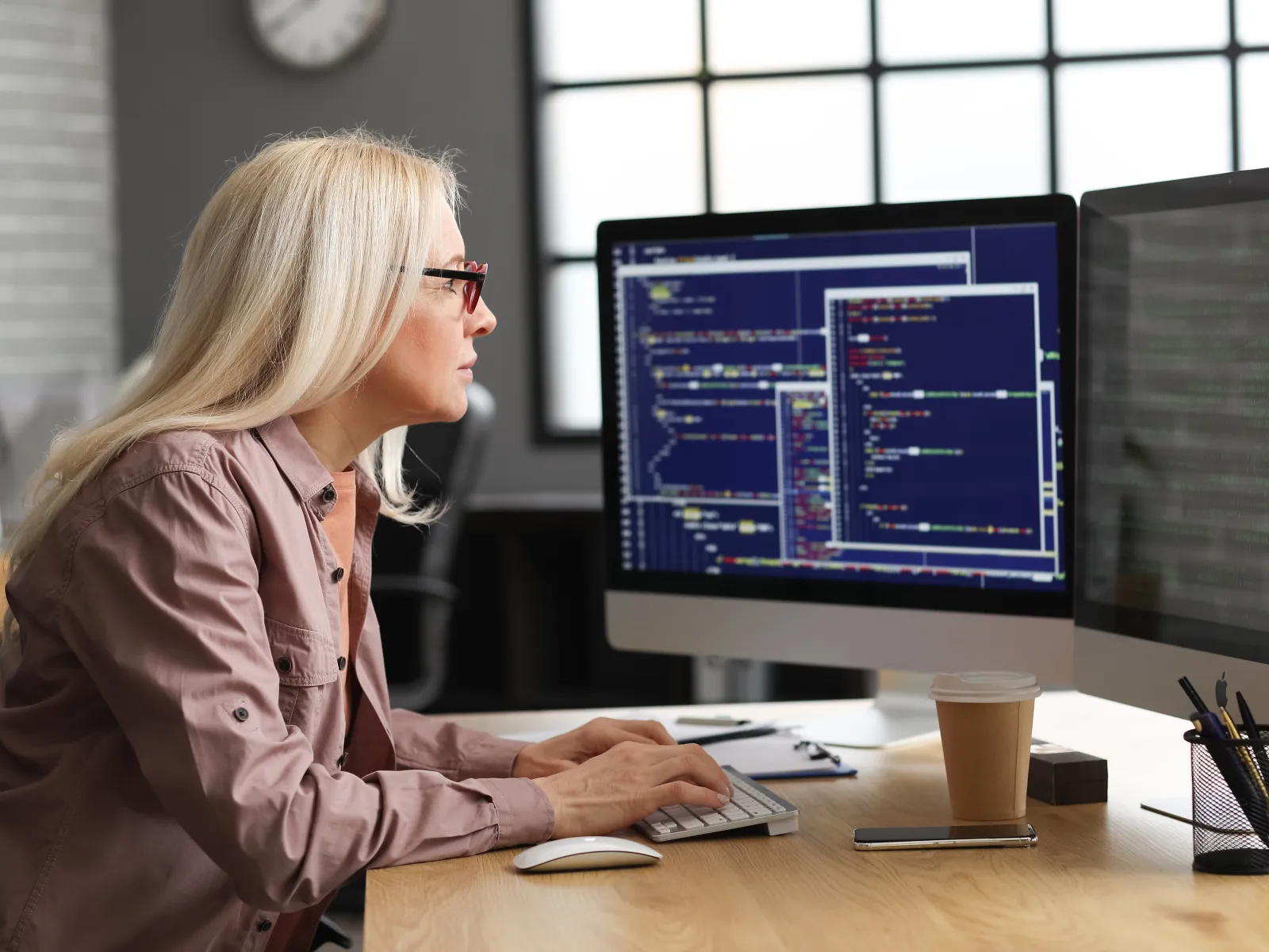 Woman coding on computer with multiple screens in a modern office setting.