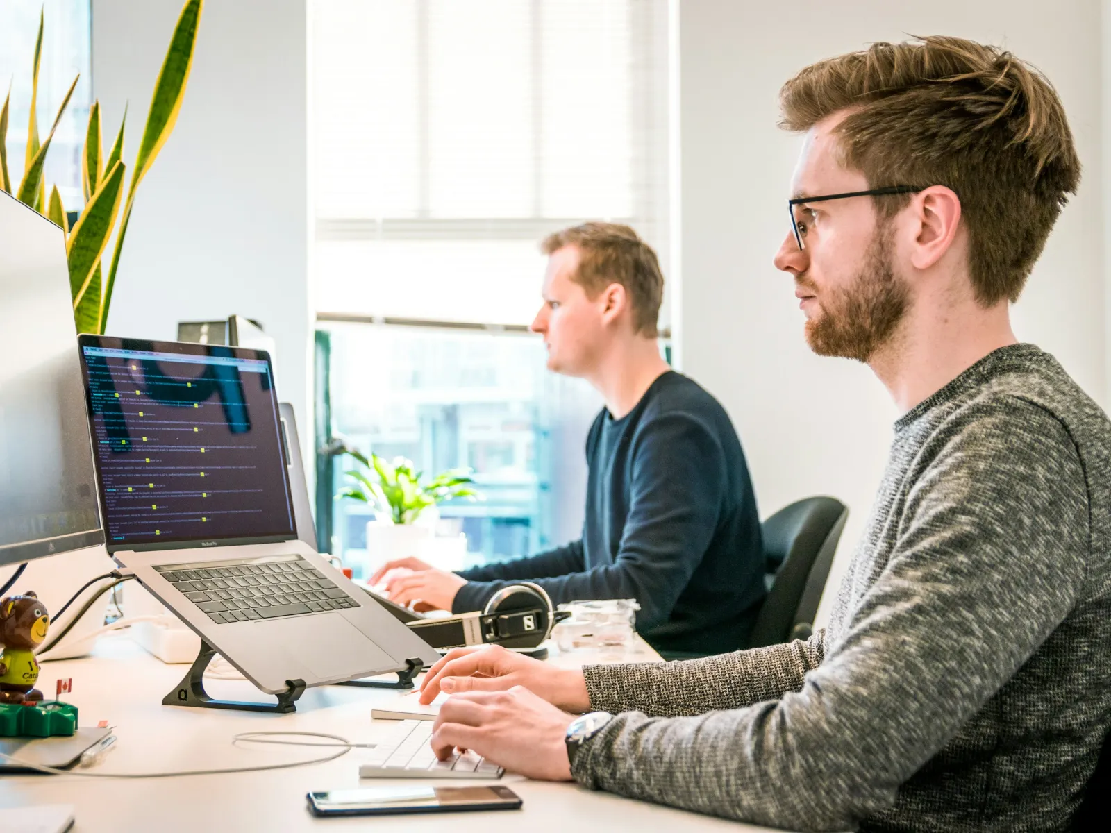 Two men focused on coding at a bright office workstation with laptops and large monitors.