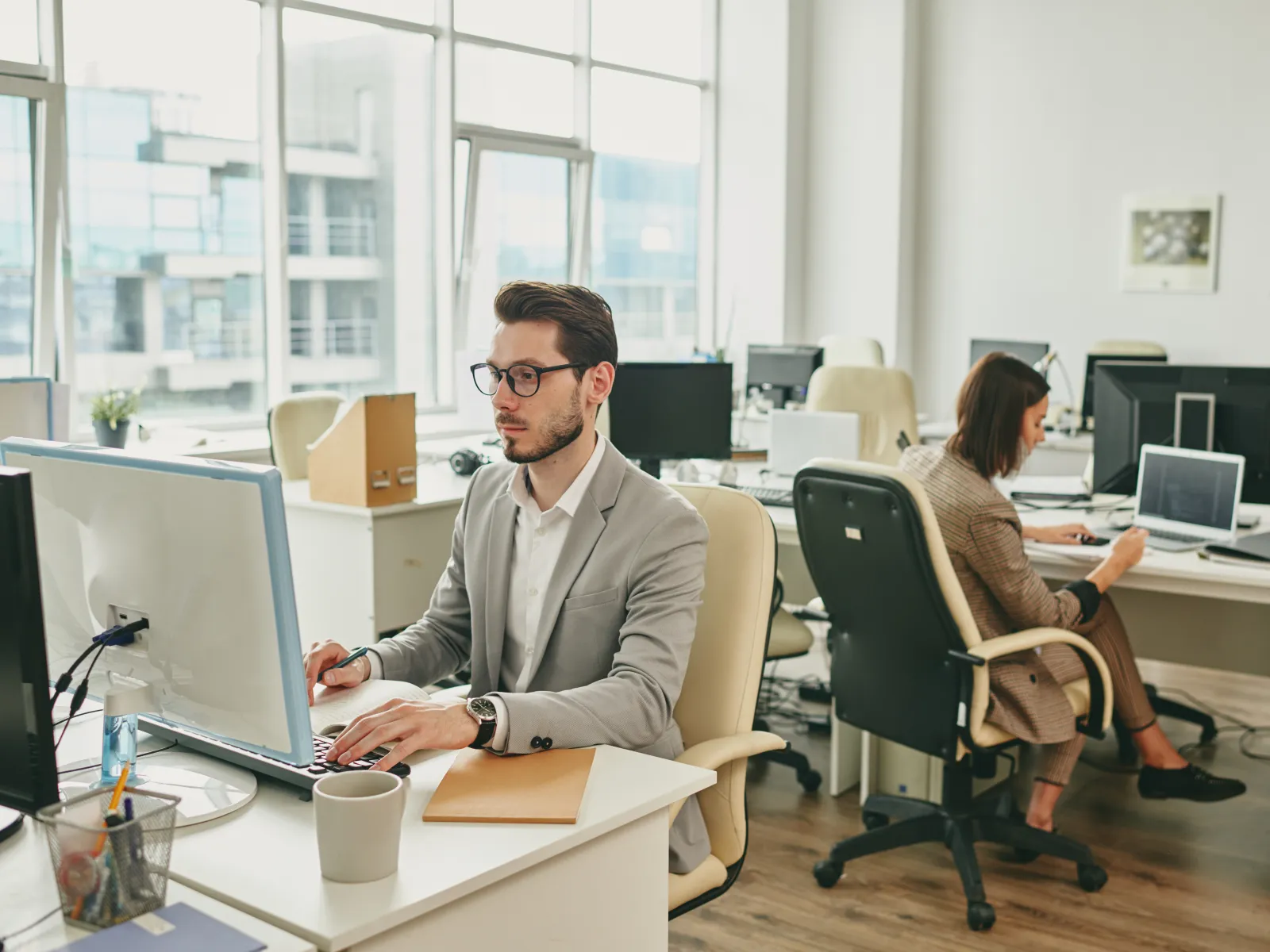 Professional office scene with a man and woman working at desks with computers and natural light from large windows.