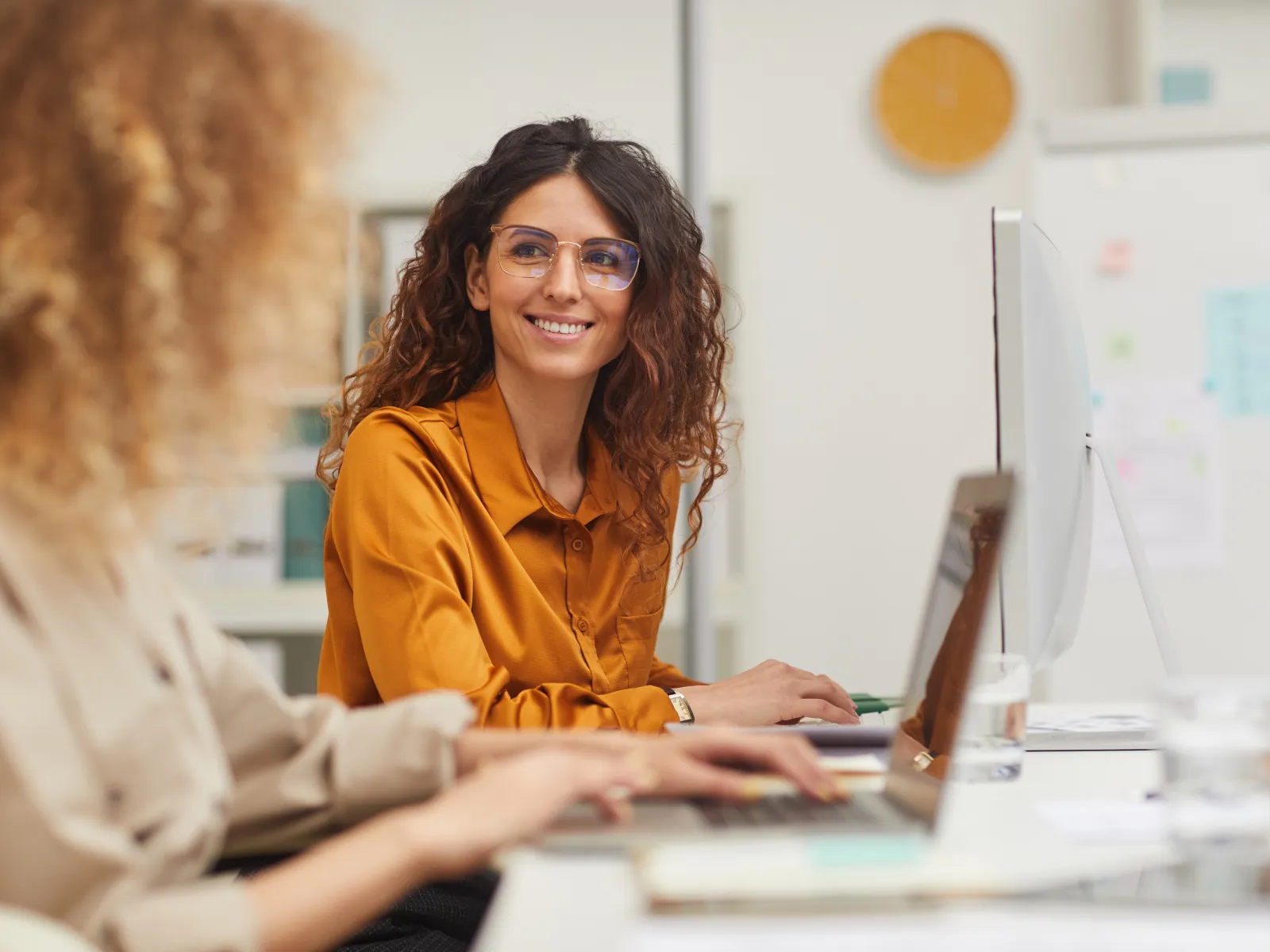 Smiling woman in orange shirt working on a computer in a modern office with a colleague nearby.