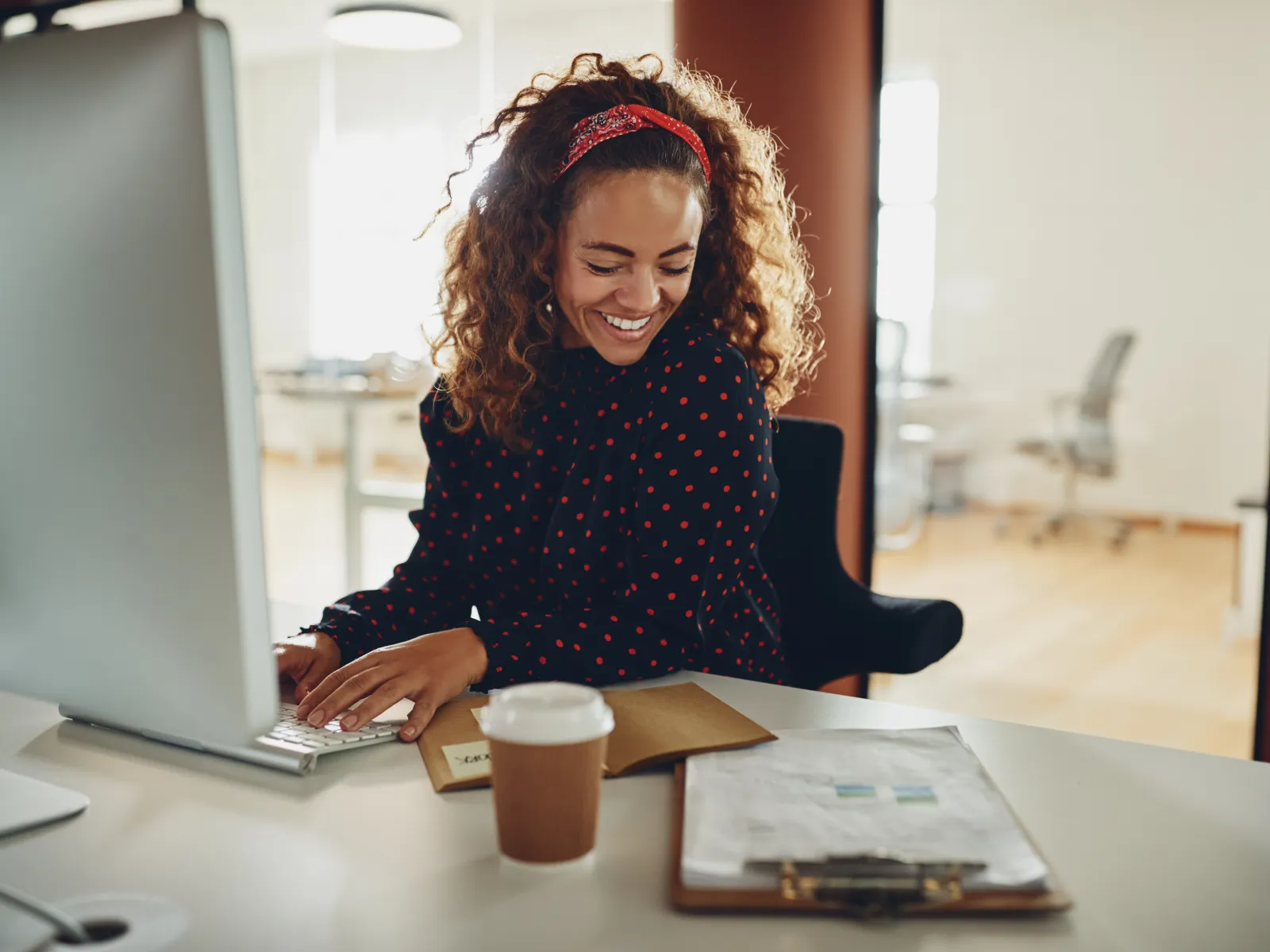 A joyful woman with curly hair smiles while working at her desk in a modern office environment.