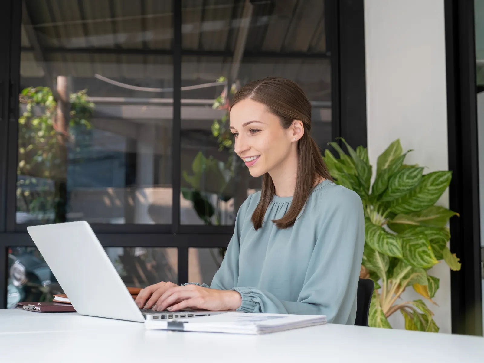 Woman working on a laptop in a modern office space with plants and large windows.