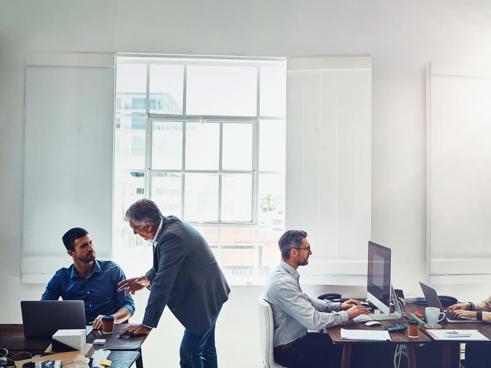 Four professionals collaborate and work on laptops and desktop computers in a bright modern office space.