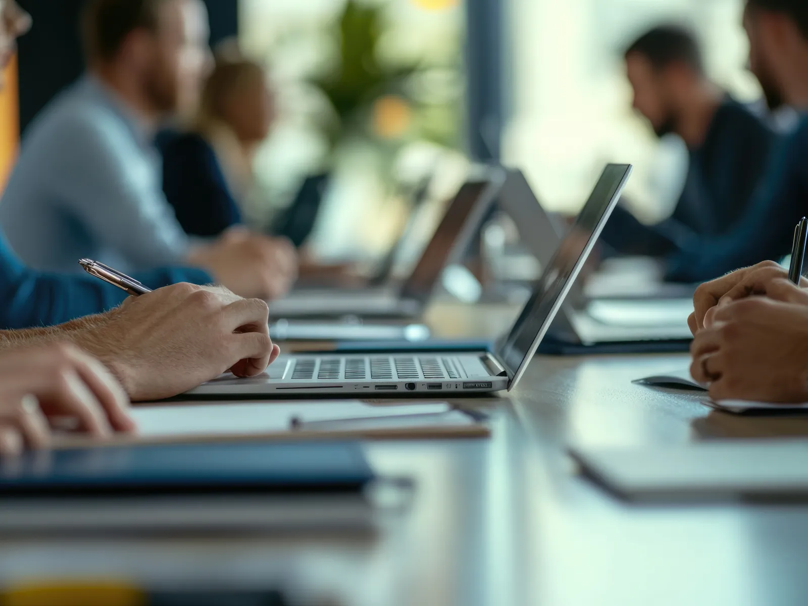 Close-up of people working on laptops and taking notes during a business meeting at a long table.