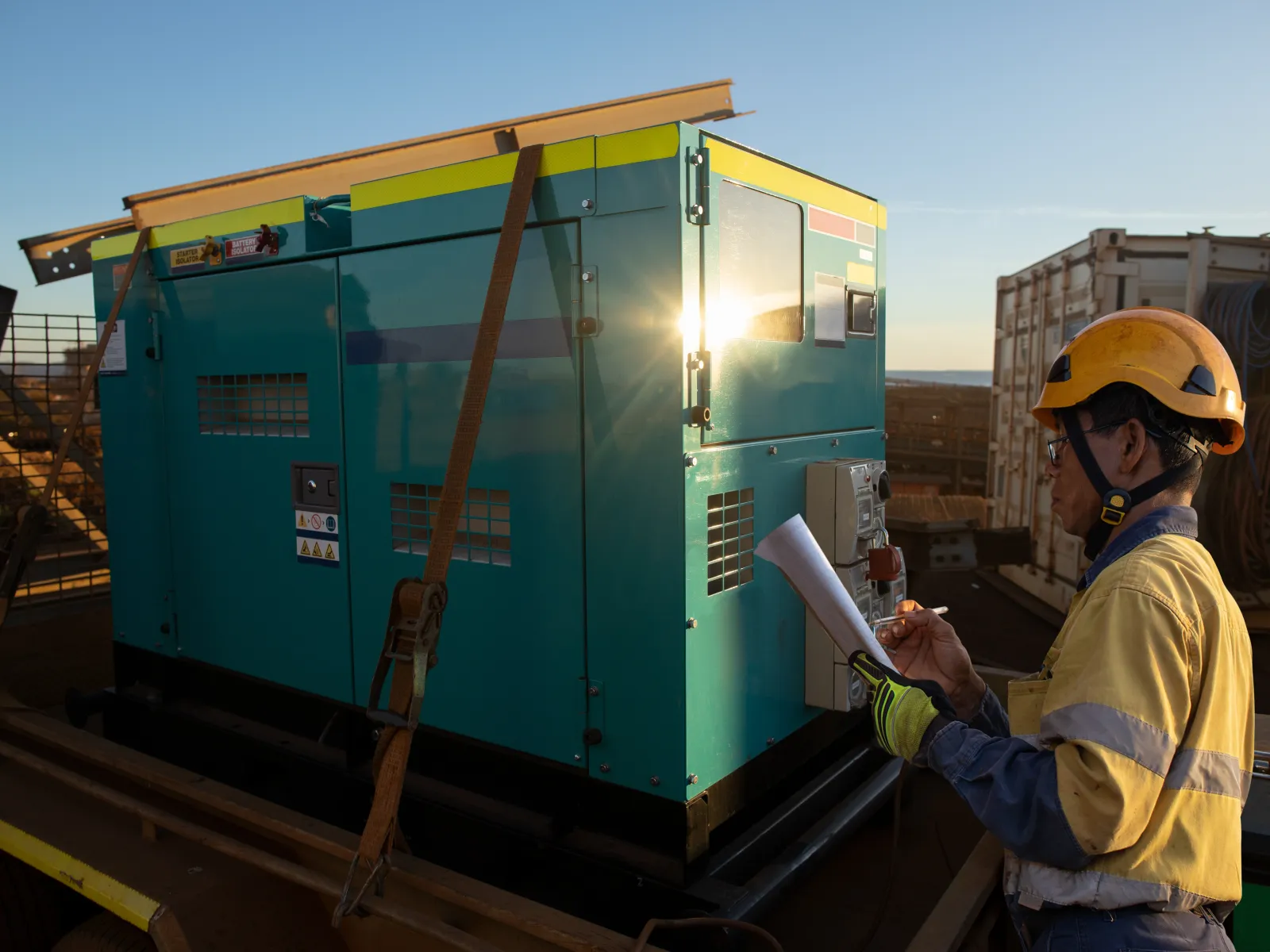 Worker reviewing details near a teal generator on a construction site at sunset.