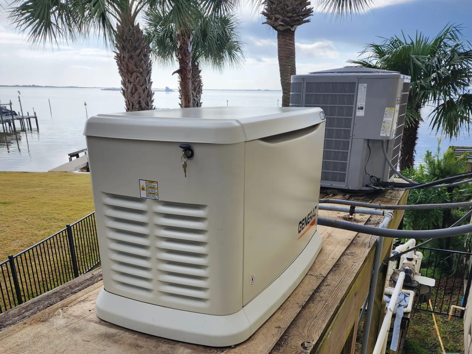Outdoor backup generator installed on wooden platform near air conditioning unit with palm trees in background