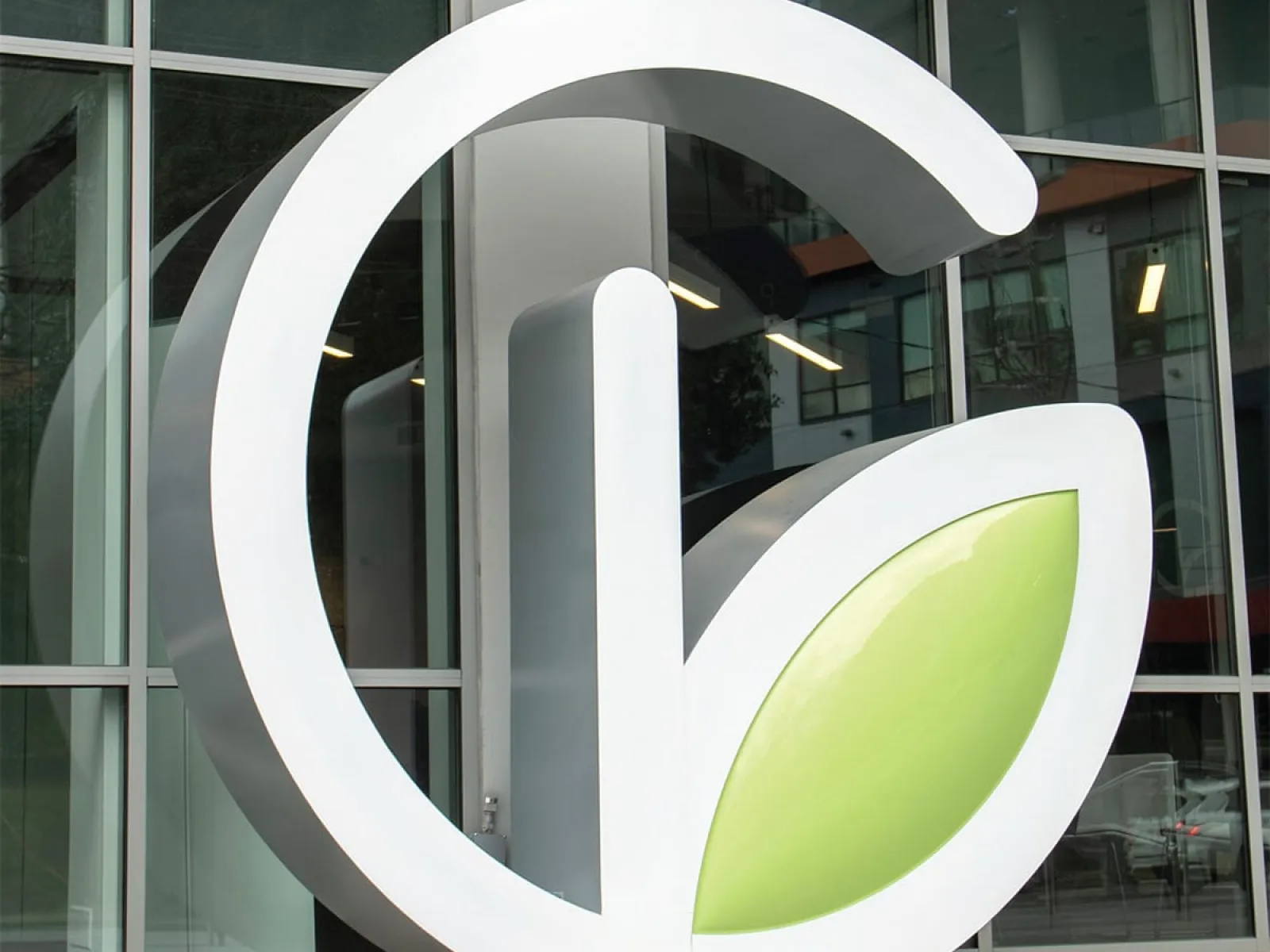 Large white G-shaped sculpture with a green leaf element outside a modern glass building.