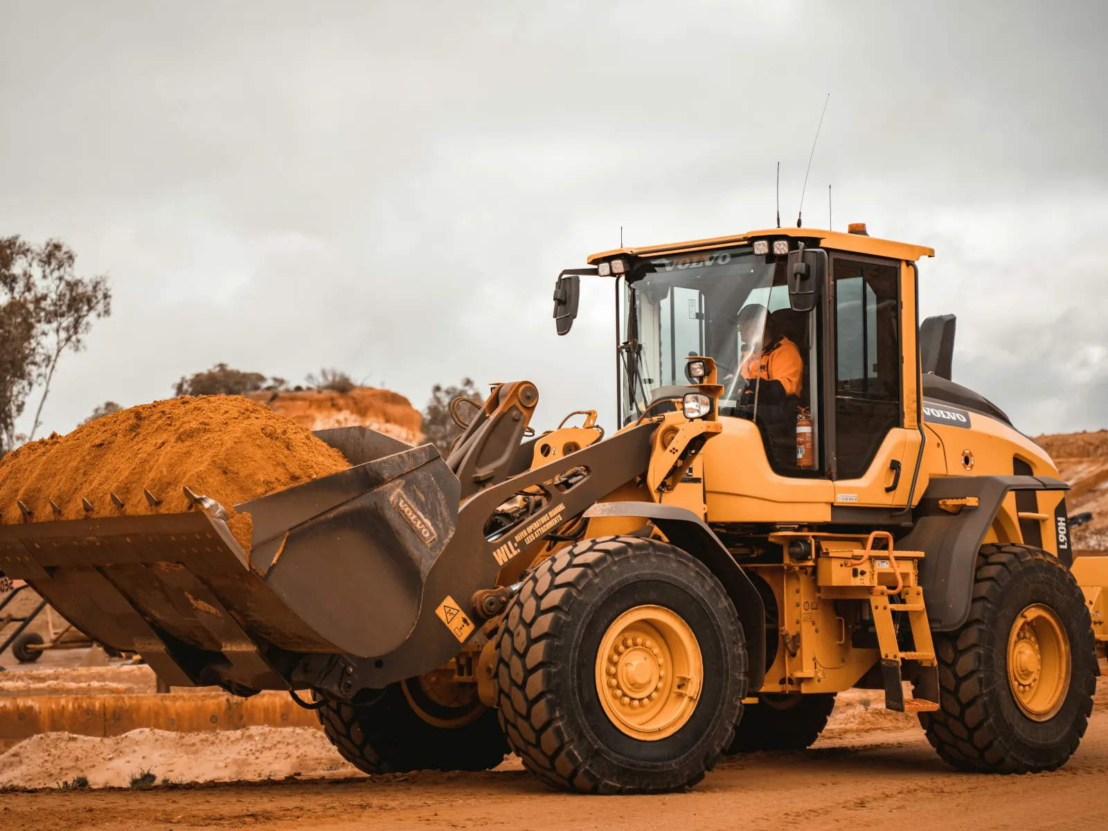 Yellow front loader excavator carrying a large bucket of dirt at a construction site under cloudy sky