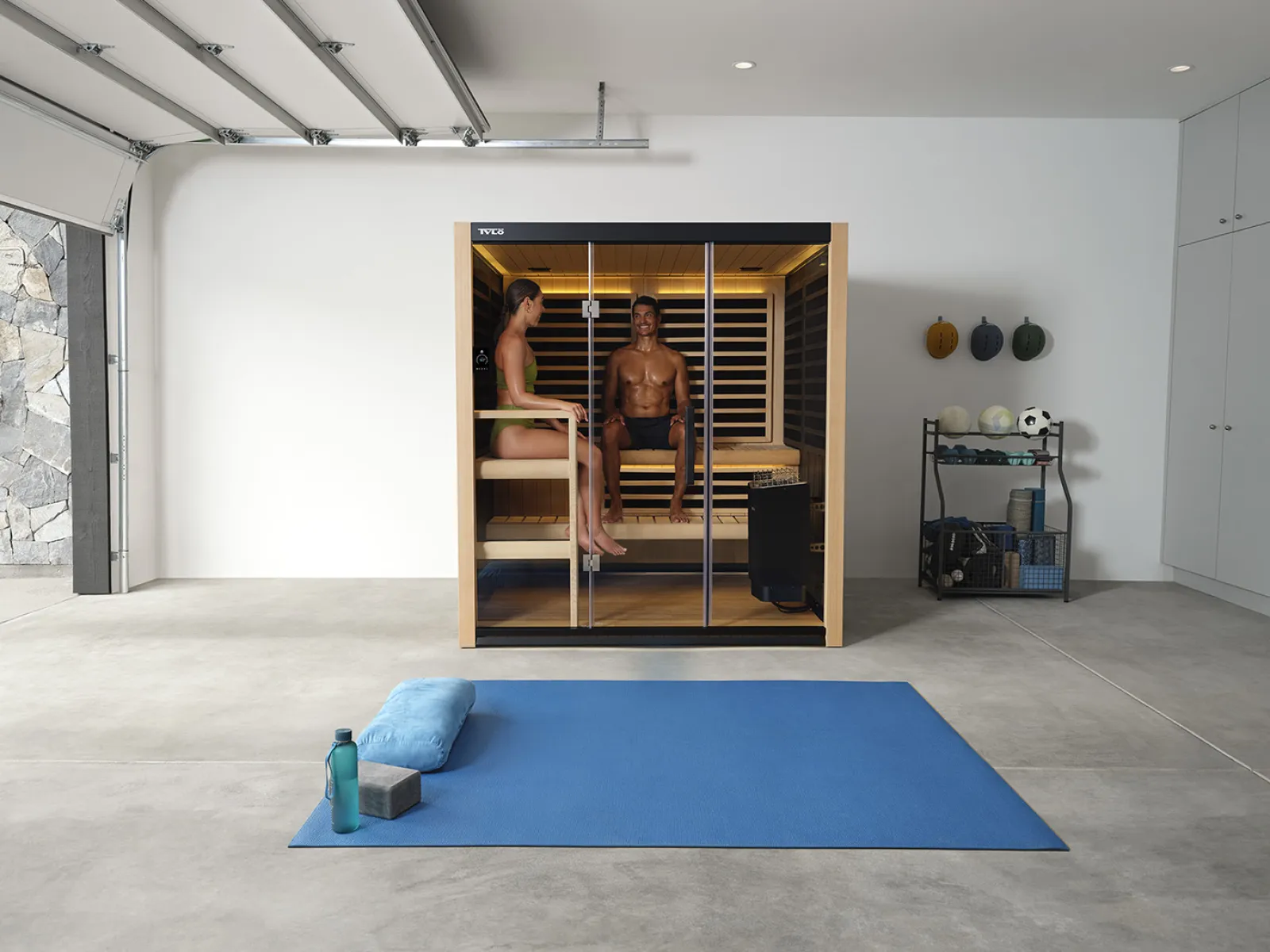Modern sauna cabin with two people inside, spacious garage gym with yoga mat, towel, and water bottle in front.