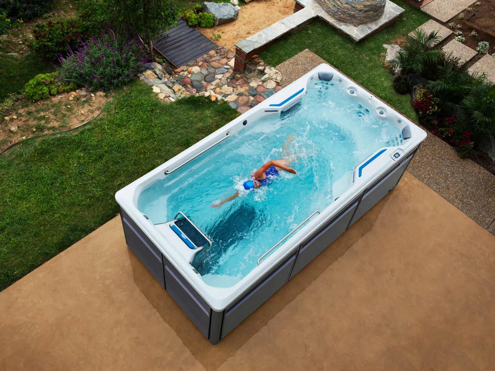 Person swimming in a backyard swim spa surrounded by greenery and stone pathways on a patio.