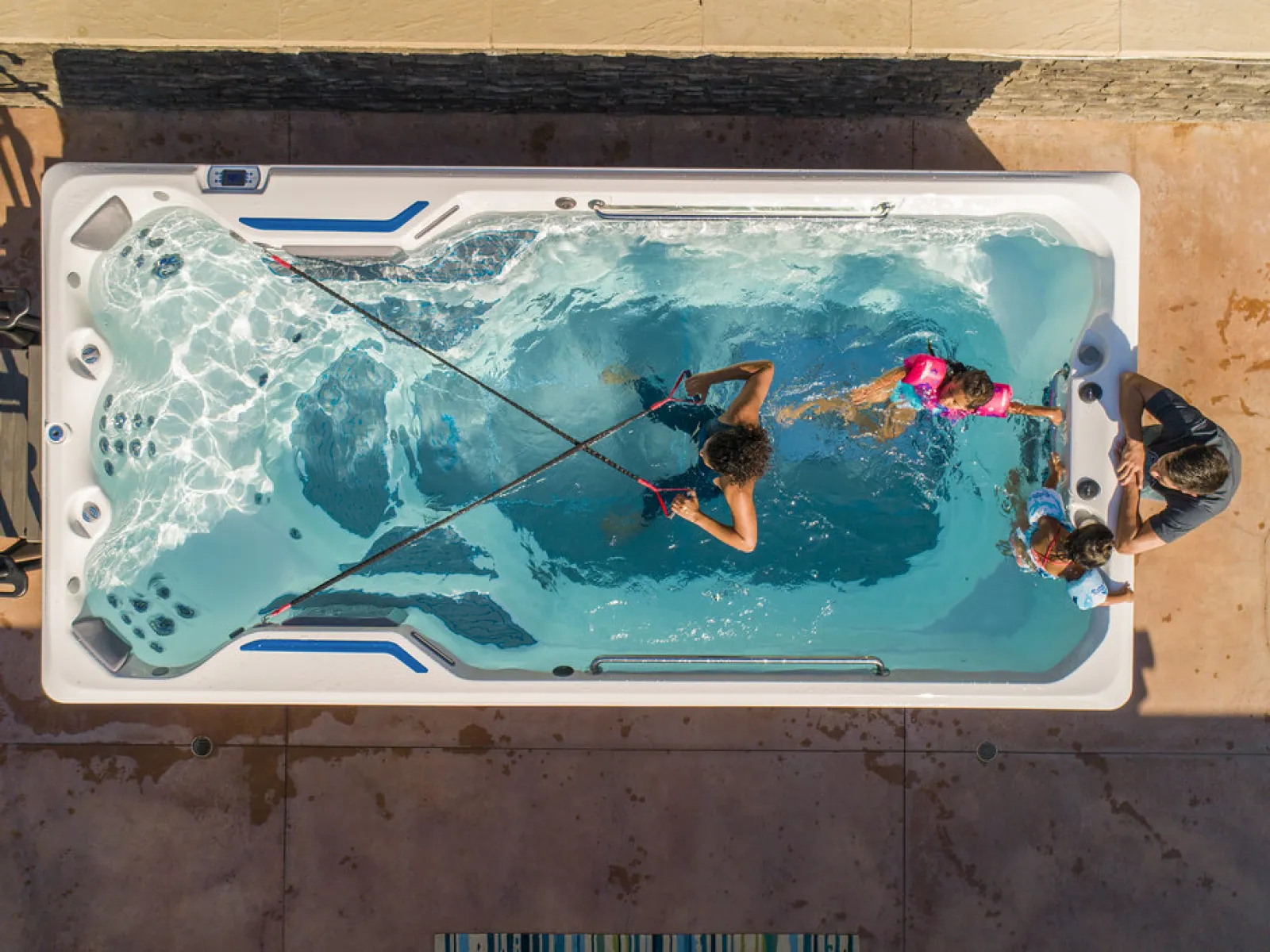 A woman swimming in a swim spa with two children and an adult supervising outside the spa on a sunny day.