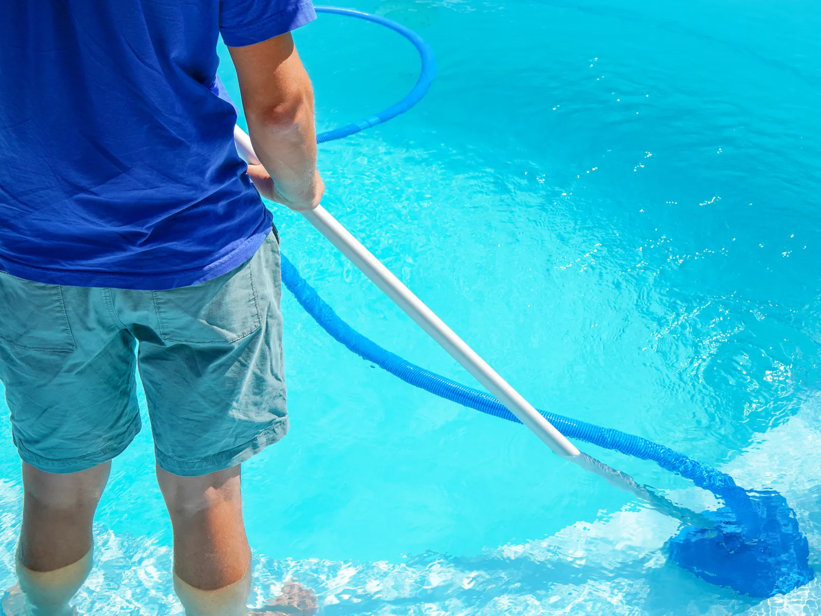 Person cleaning a bright blue swimming pool with a vacuum hose in clear water on a sunny day.
