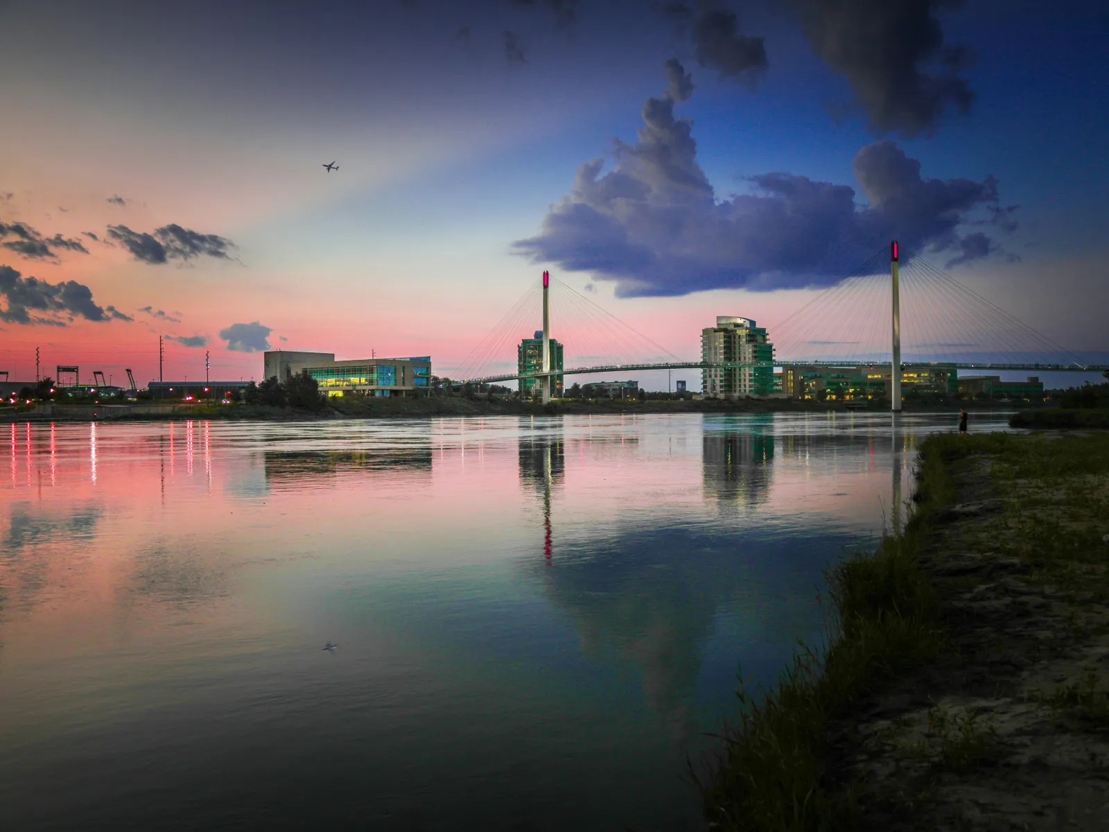 Sunset view of a calm river reflecting city buildings, a cable-stayed bridge, and a plane in the sky.