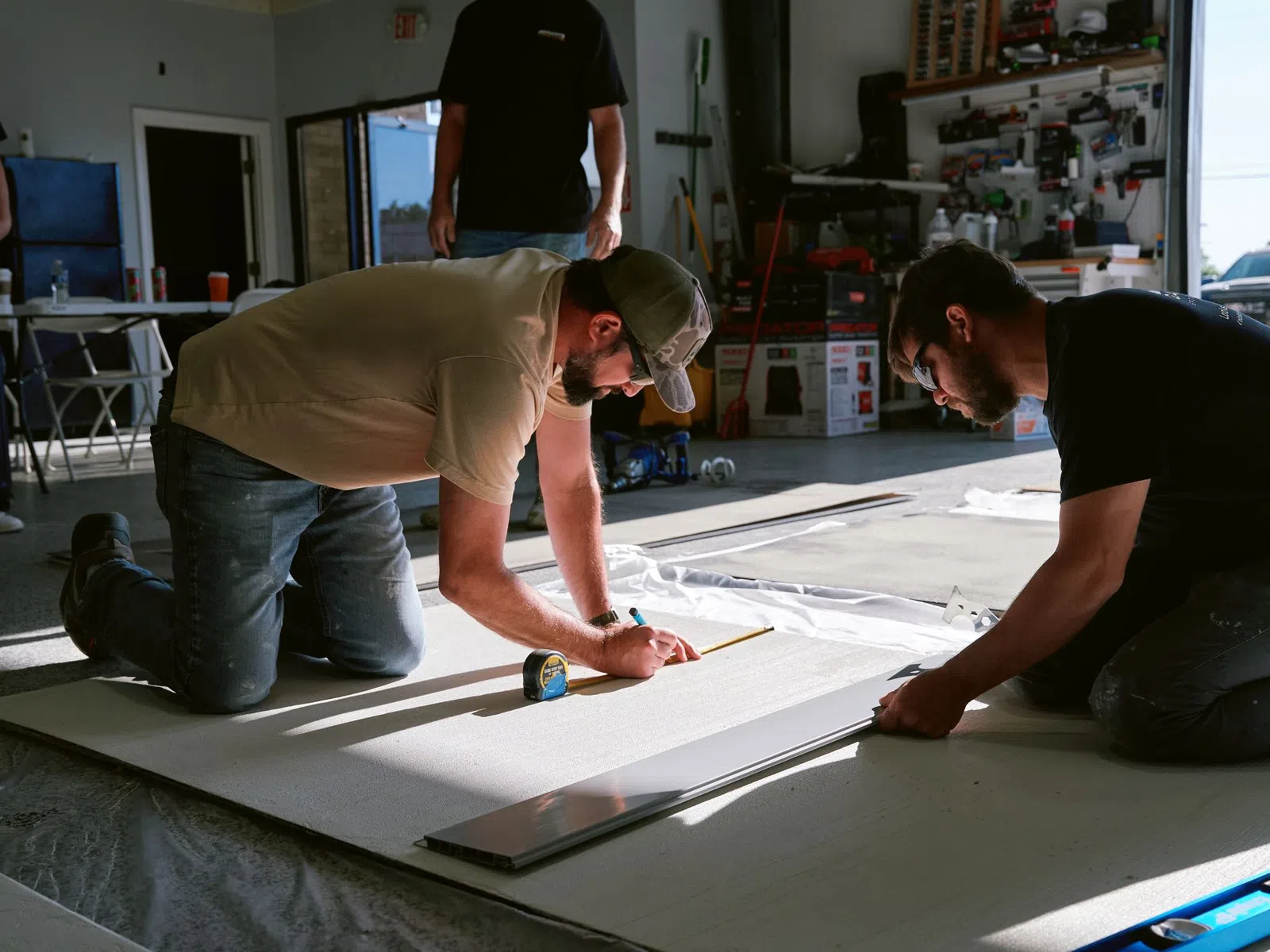 Two men kneeling and measuring a large board in a workshop with tools and equipment around them.