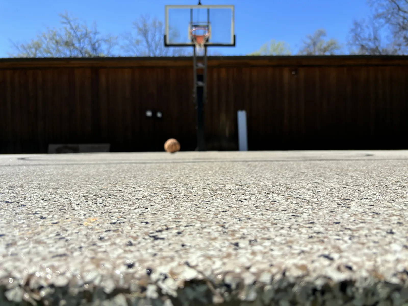 Close-up of textured concrete basketball court surface with hoop and ball near a wooden fence under clear sky.