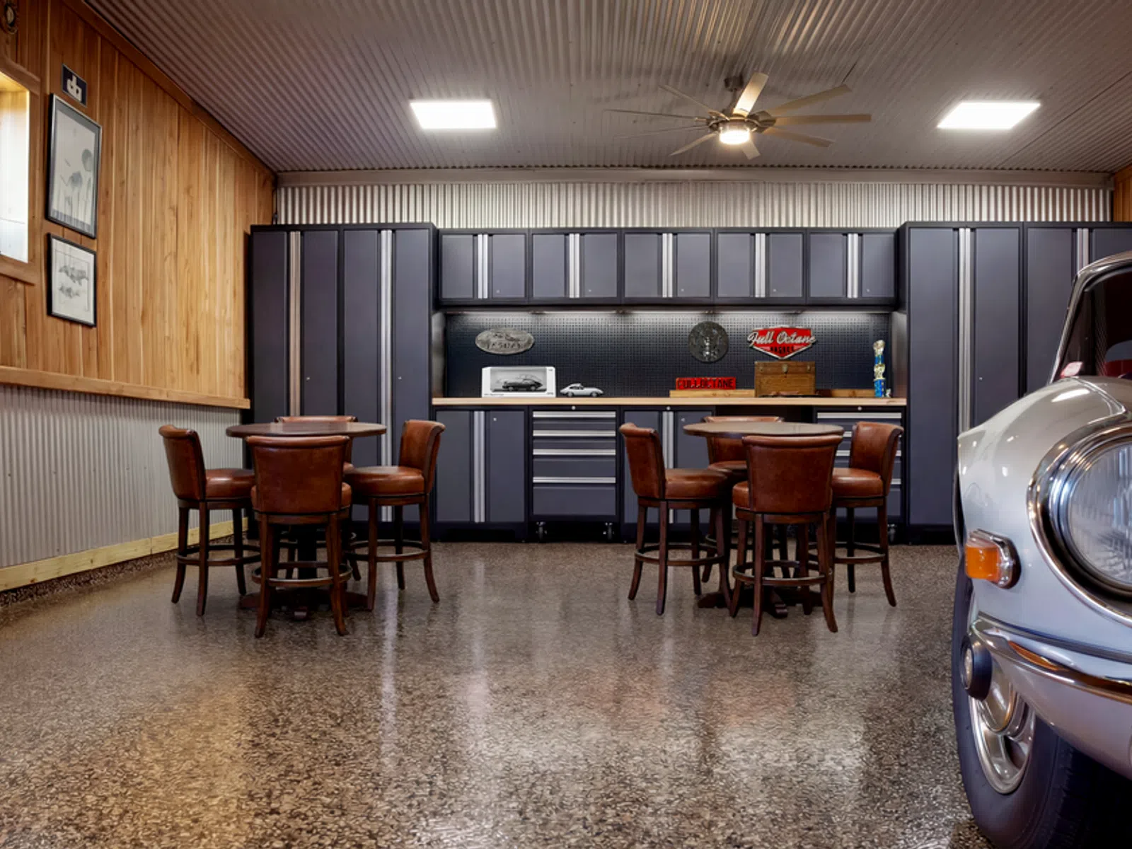 Retro-style garage interior with vintage car, wooden tables, leather chairs, and metal cabinets under ceiling fans.