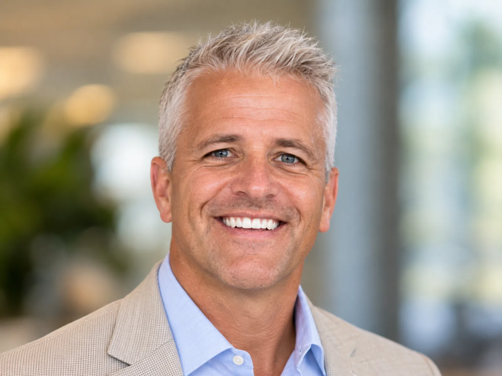 Smiling middle-aged man with gray hair wearing a beige blazer and light blue shirt in an office setting