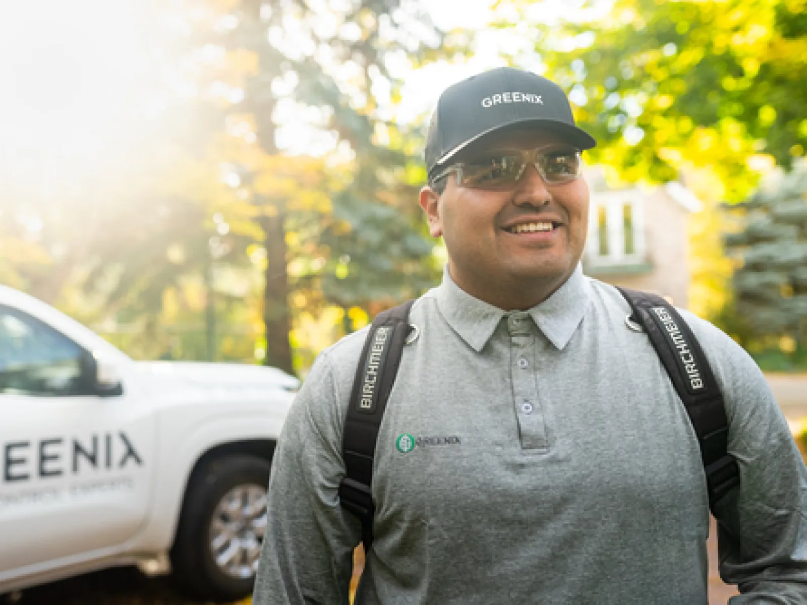 Smiling pest control technician wearing Greenix uniform and cap stands outdoors near Greenix service vehicle