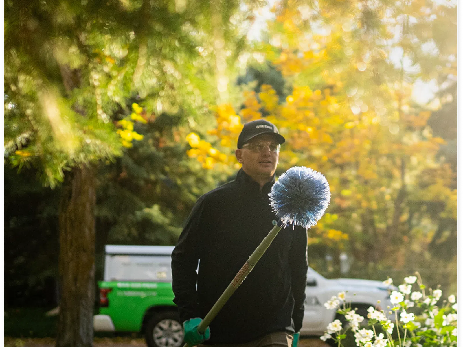 Man in black jacket and cap holding a long-handled blue brush in a sunlit garden with yellow and white flowers.